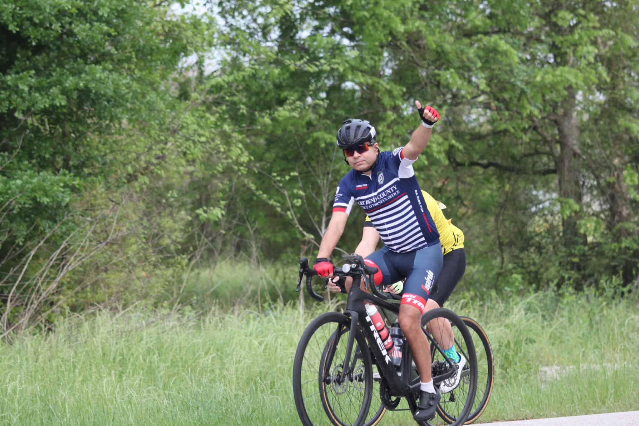 Vibrant cyclist in Bluebonnet Express bike jersey riding through lush green forest, raising thumb up, enjoying outdoor cycling adventure.