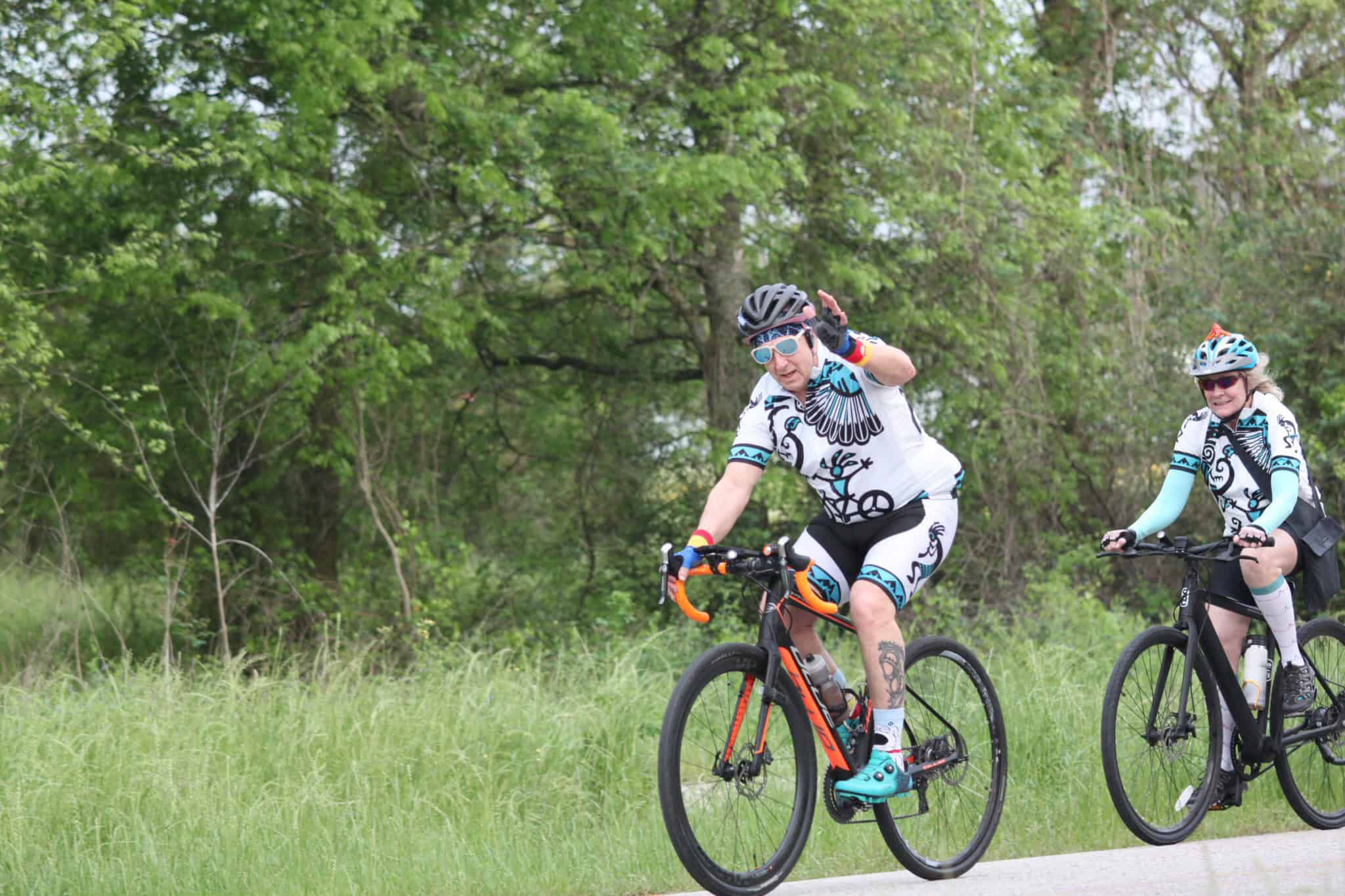 Brightly dressed cyclists riding mountain bikes on lush green trail with trees in the background.
