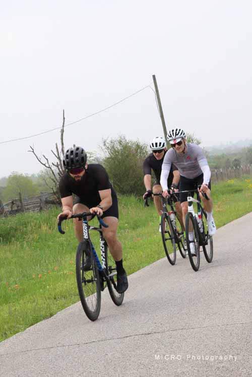 Velo cyclists riding on scenic rural bike trail with greenery and overcast sky.