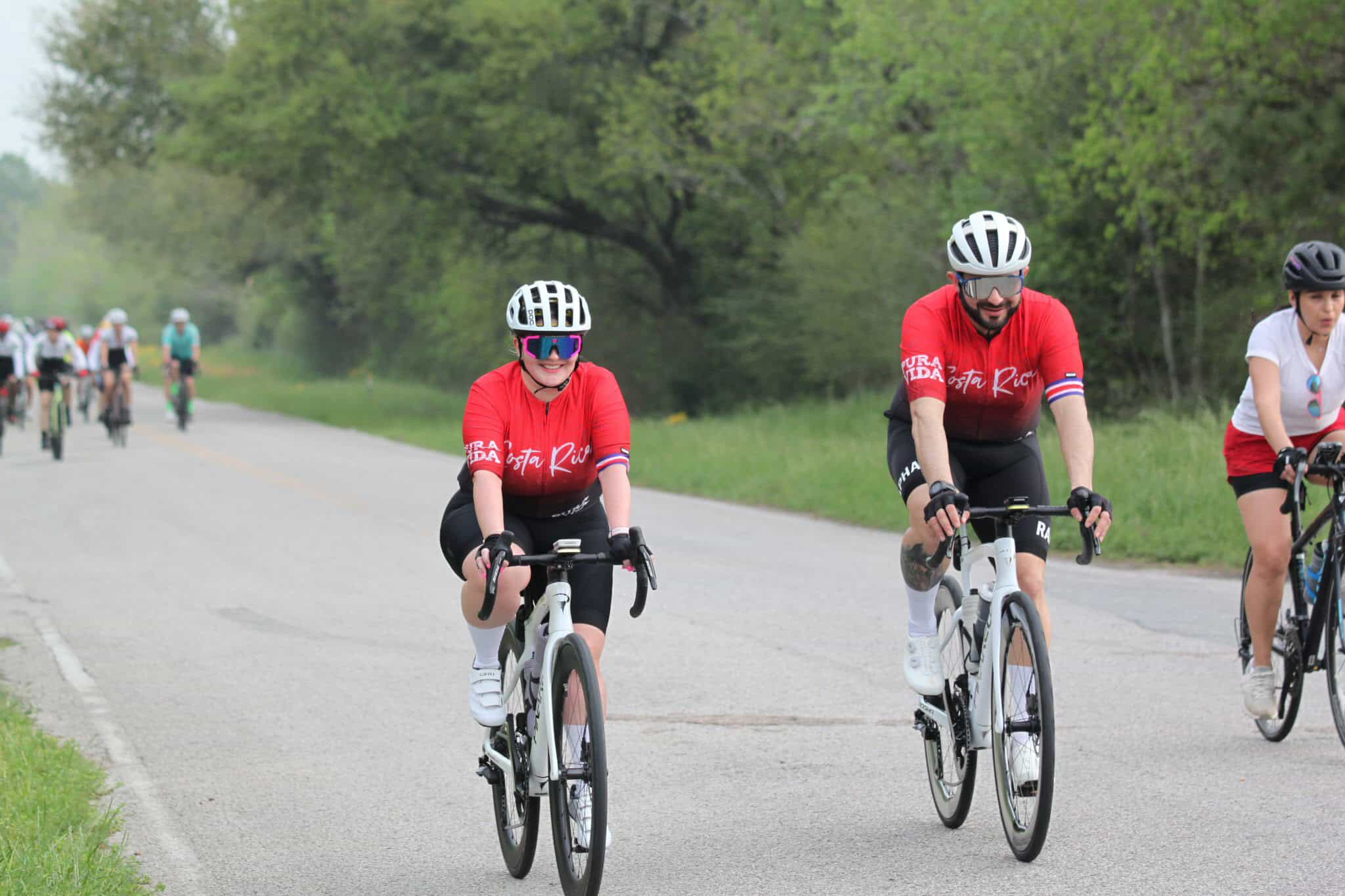 Bright female cyclist wearing a red jersey and helmet, smiling while riding on scenic countryside road.