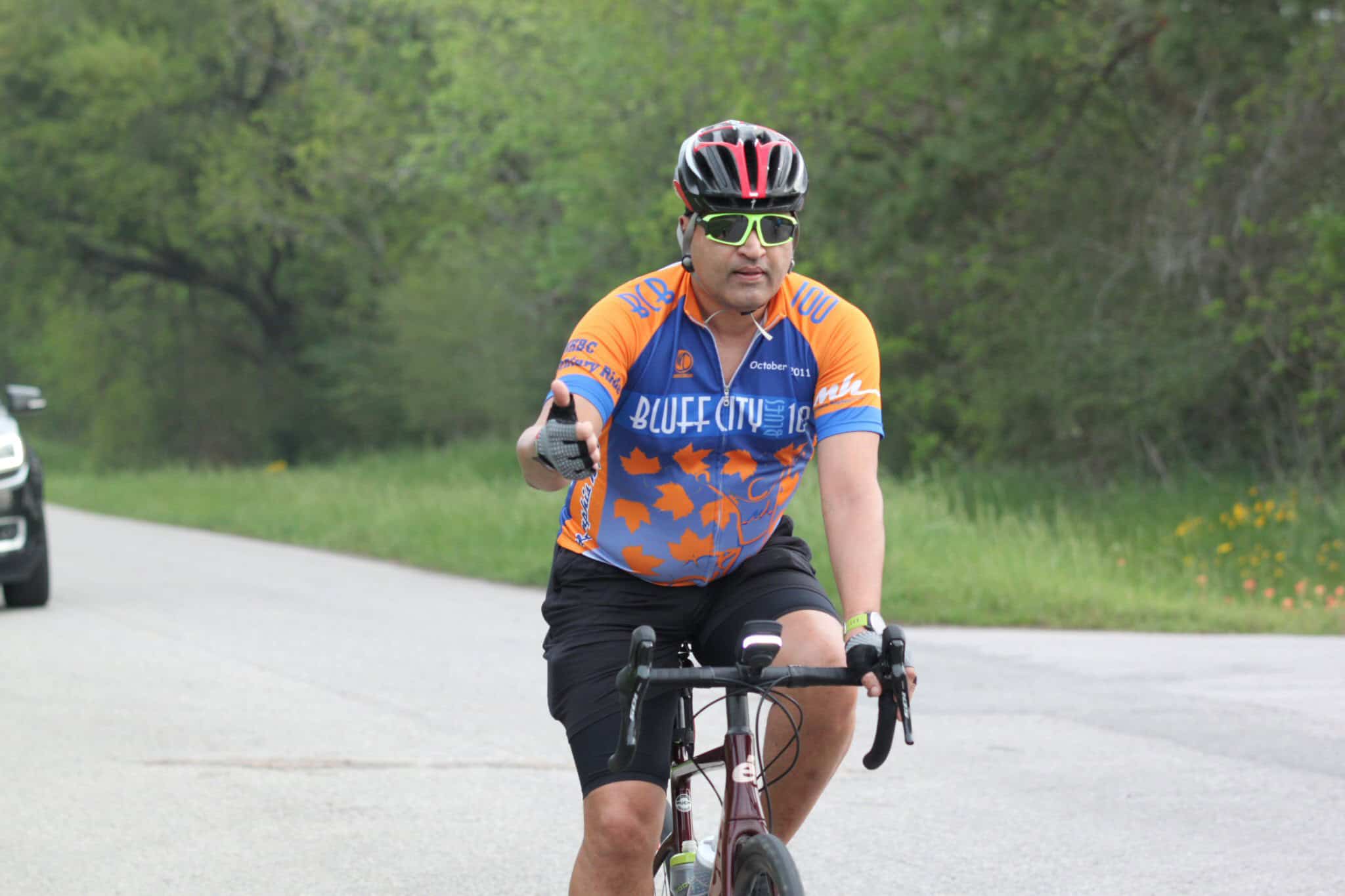 Bike rider in Orange and Blue Buff City cycling jersey on scenic road with greenery.