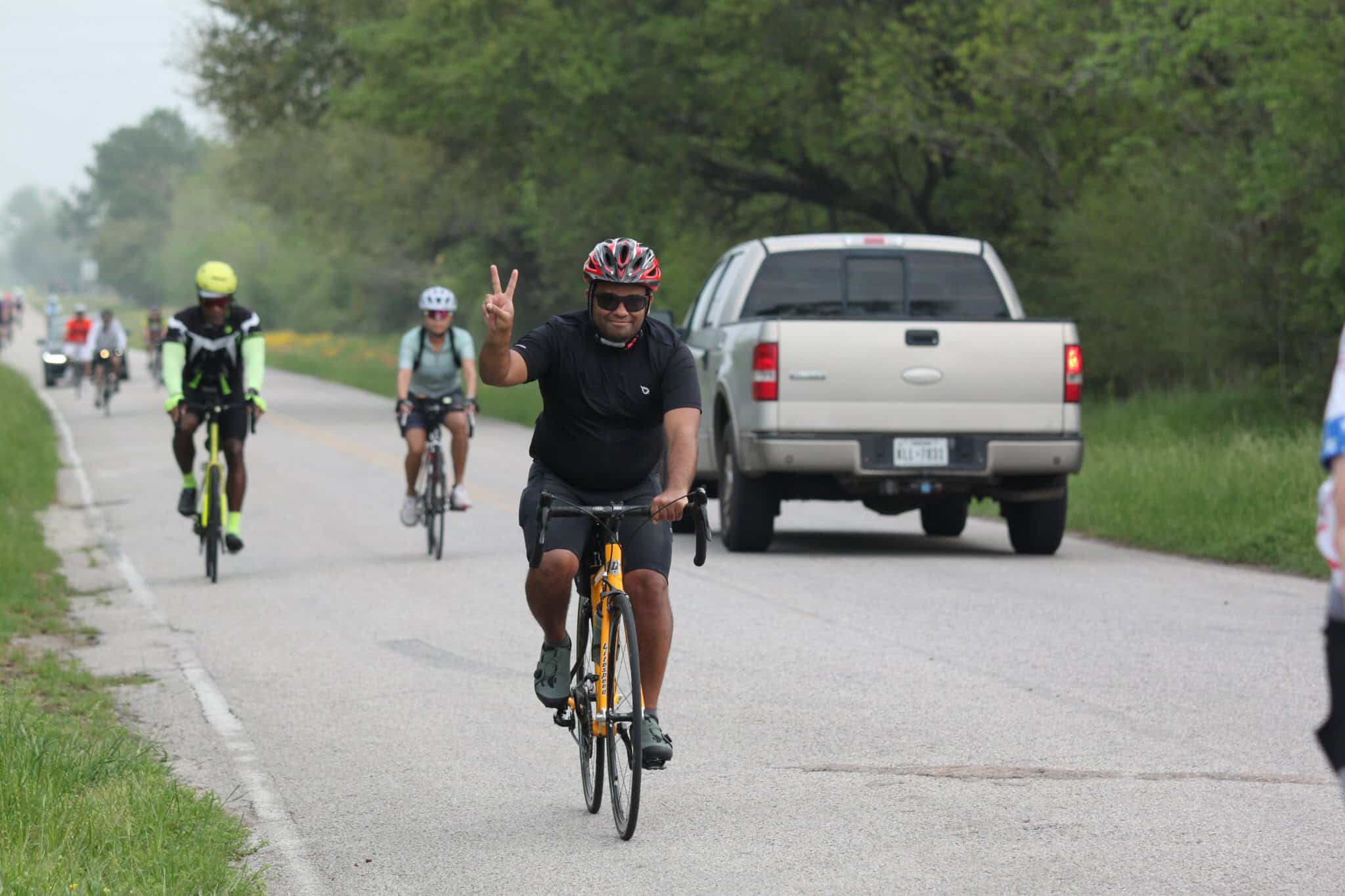 Bicycle rider making peace sign during group cycling event on scenic countryside road.