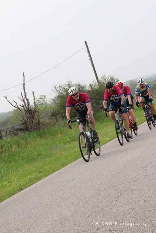 Vélo group riding on a country road wearing blue and pink jerseys, promoting Bluebonnet Express cycling event.