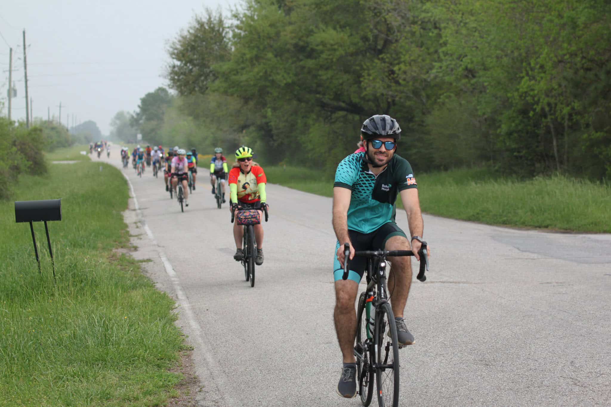 Elegant cyclists participating in Bluebonnet Express cycling event on scenic rural road with lush green trees.