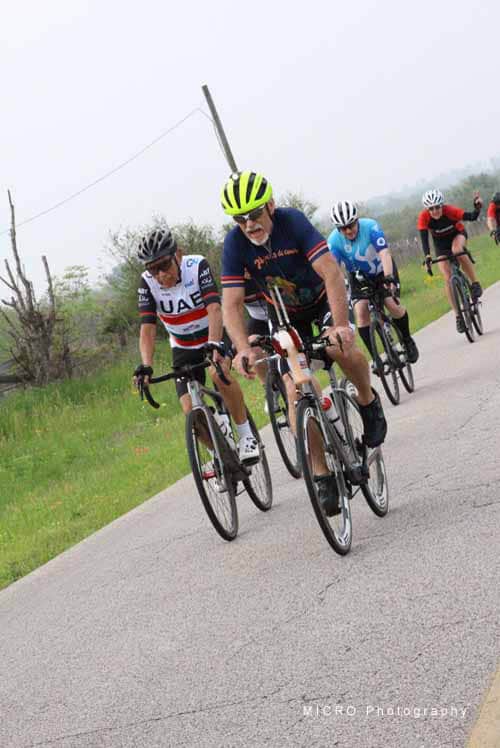 Riders in Bluebonnet Express cycling event on a scenic rural road, wearing colorful jerseys and helmets, supporting community cycling races.