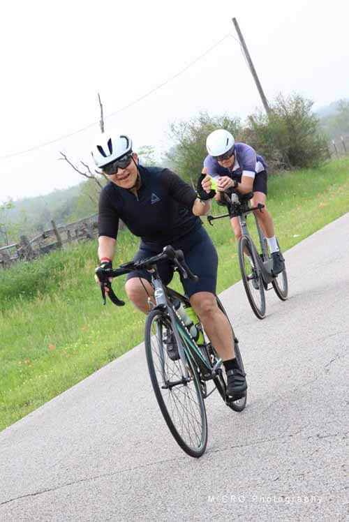 1. Two cyclists riding on a scenic rural road during daytime, wearing helmets and casual athletic gear.