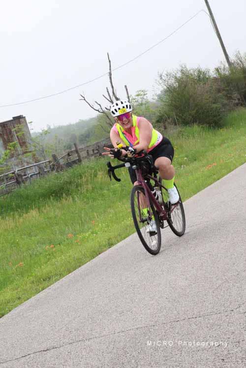 Bright cyclist wearing a helmet with a tree branch on her head riding on the road.