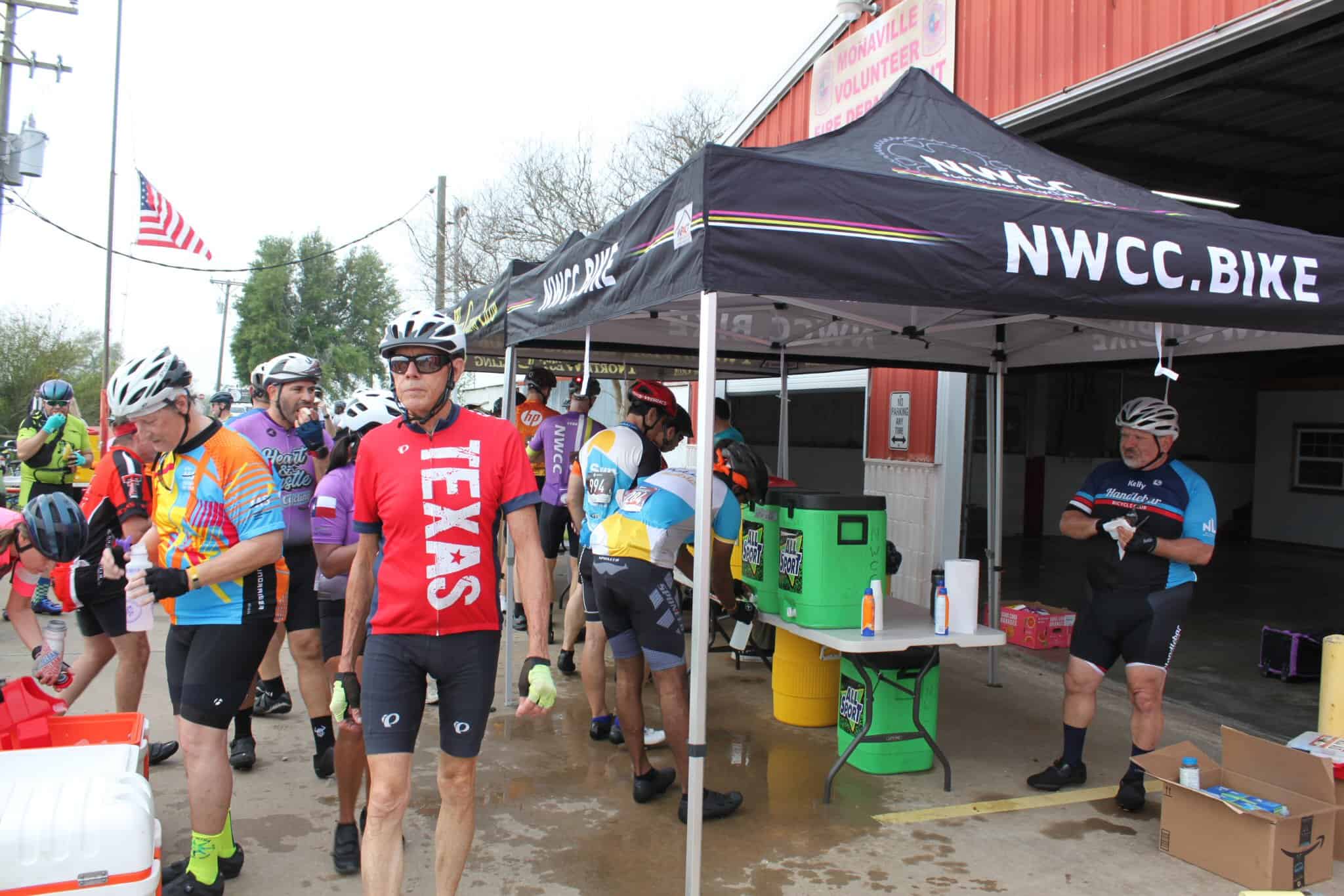 Cyclists at Bluebonnet Express event under NWCC.BIKE tent, team biking gear, hydration station setup, outdoor cycling race scene.