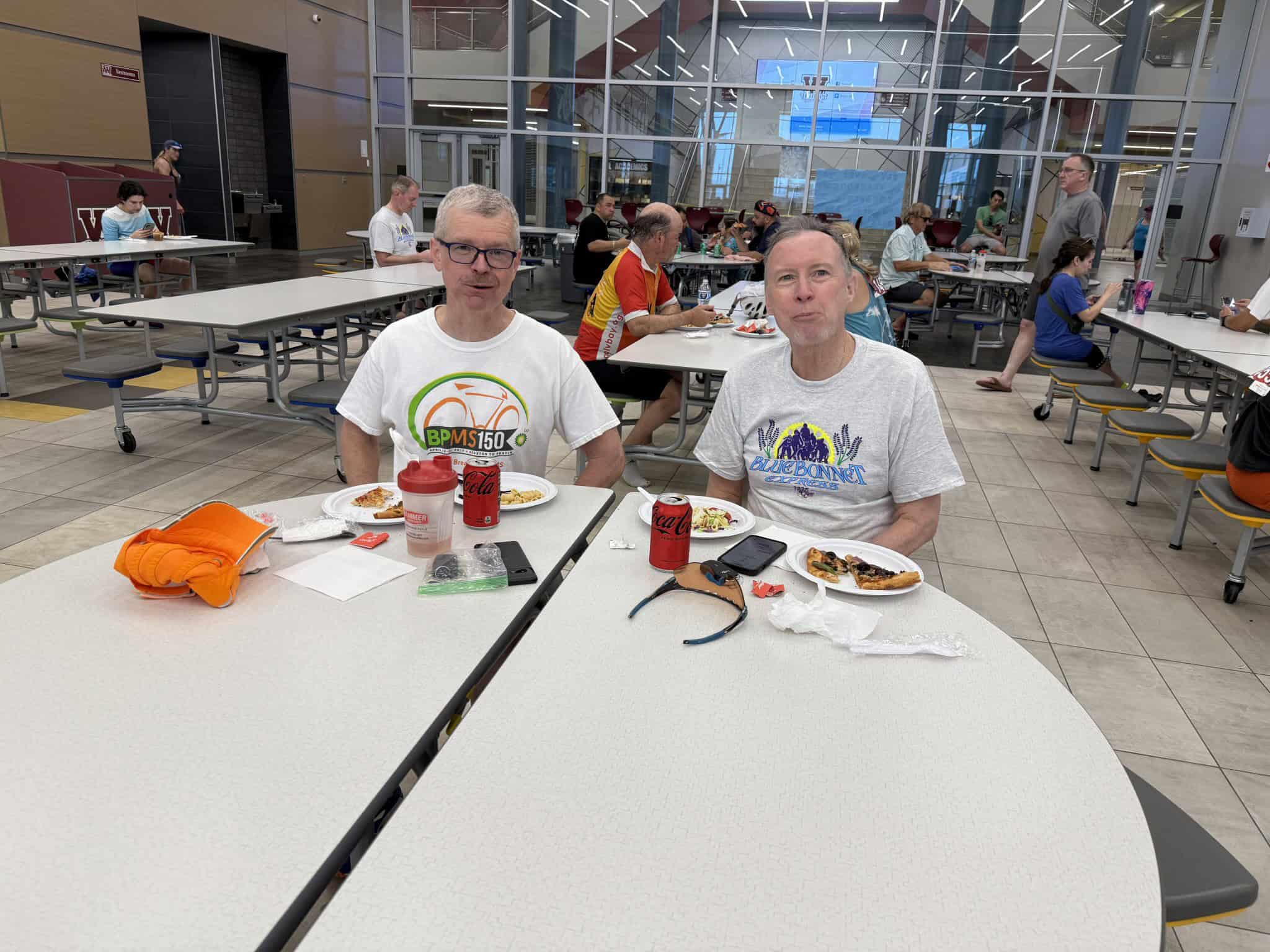 Comfortable indoor cafeteria with two bikers enjoying meals and drinks, surrounded by fellow cyclists and travelers.