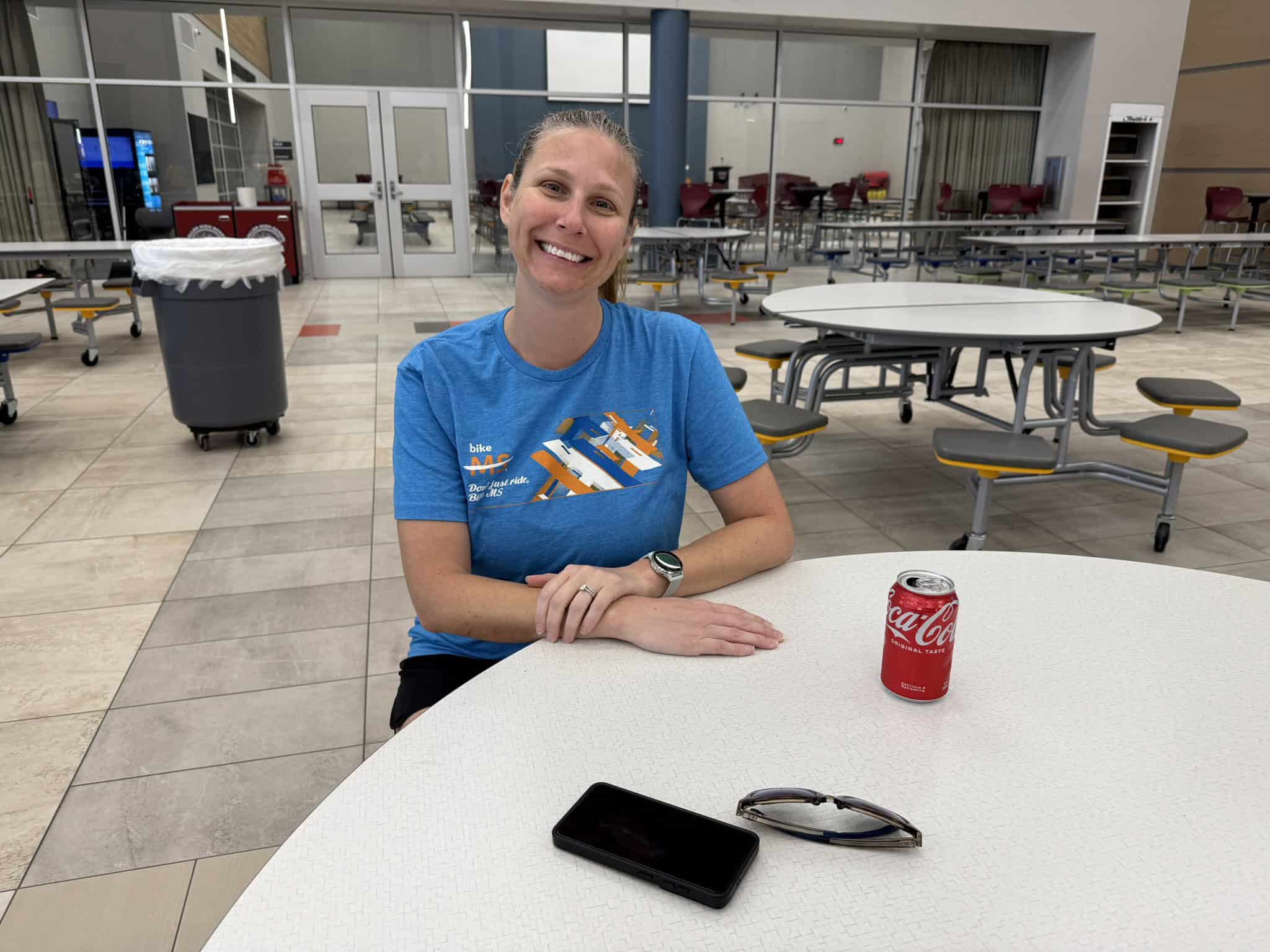 Bright smiling woman in blue Bicycle MS t-shirt sitting at a cafeteria table with Coca-Cola can and sunglasses, indoor dining area.