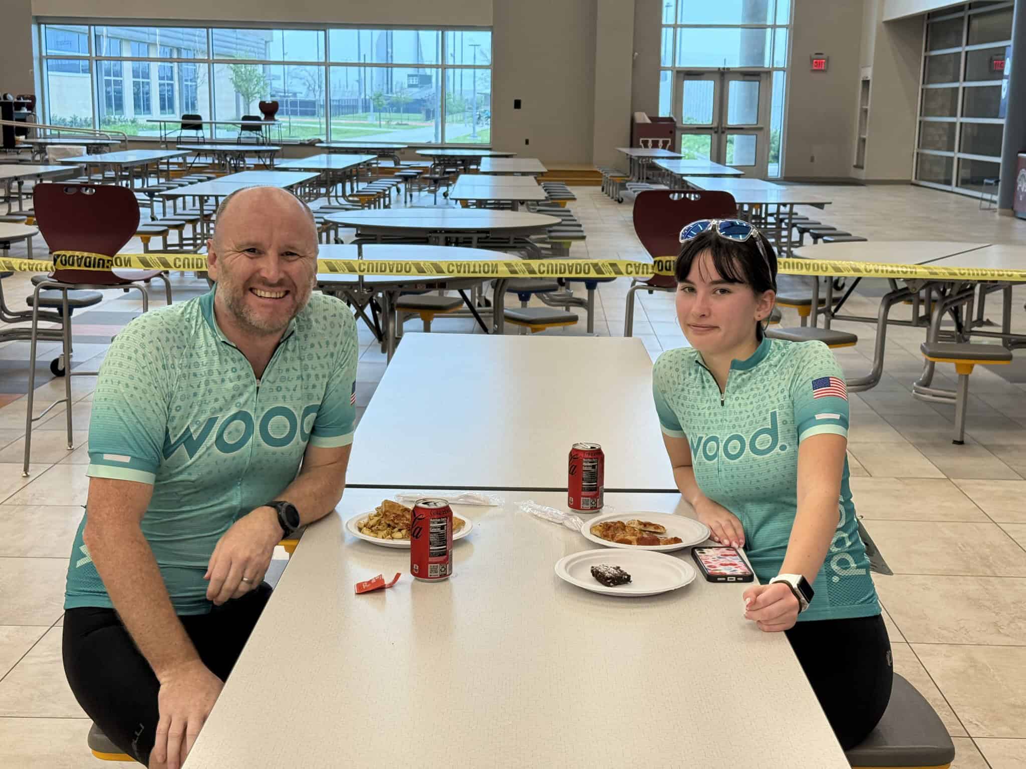Cheerful cyclists enjoying a meal at a community event, wearing matching Wood Cycling Jerseys, inside a spacious hall with COVID caution tape.