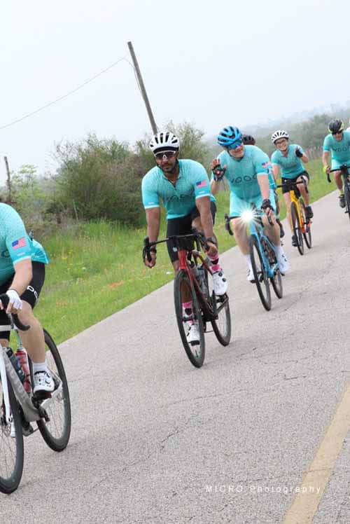 Bright blue jerseys, cyclists riding on rural road, Bluebonnet Express, group cycling event, outdoor biking, team ride, Texas scenic route.