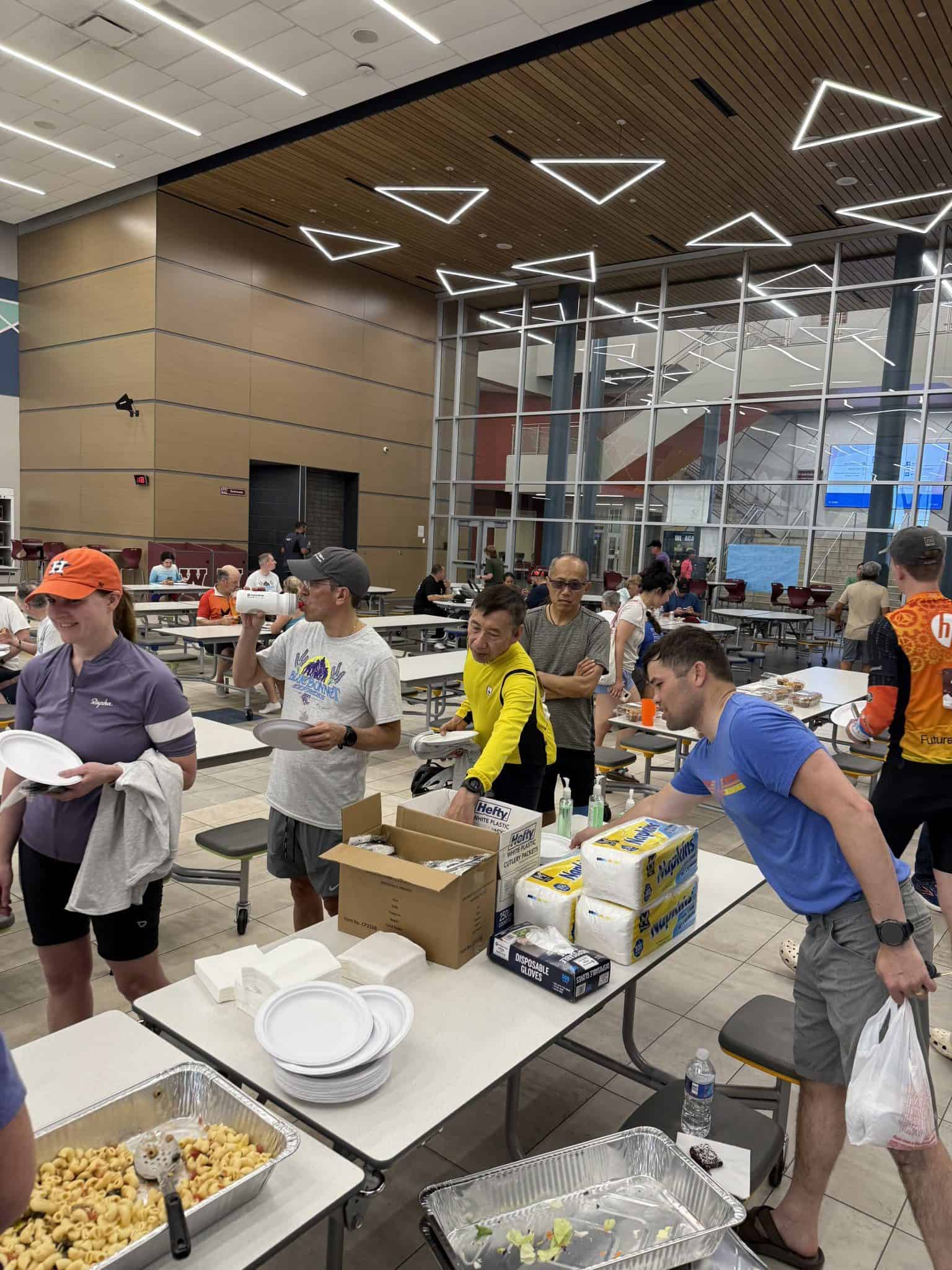 Bright indoor dining hall with diverse people volunteering at food distribution table. Modern ceiling lights, large glass windows.