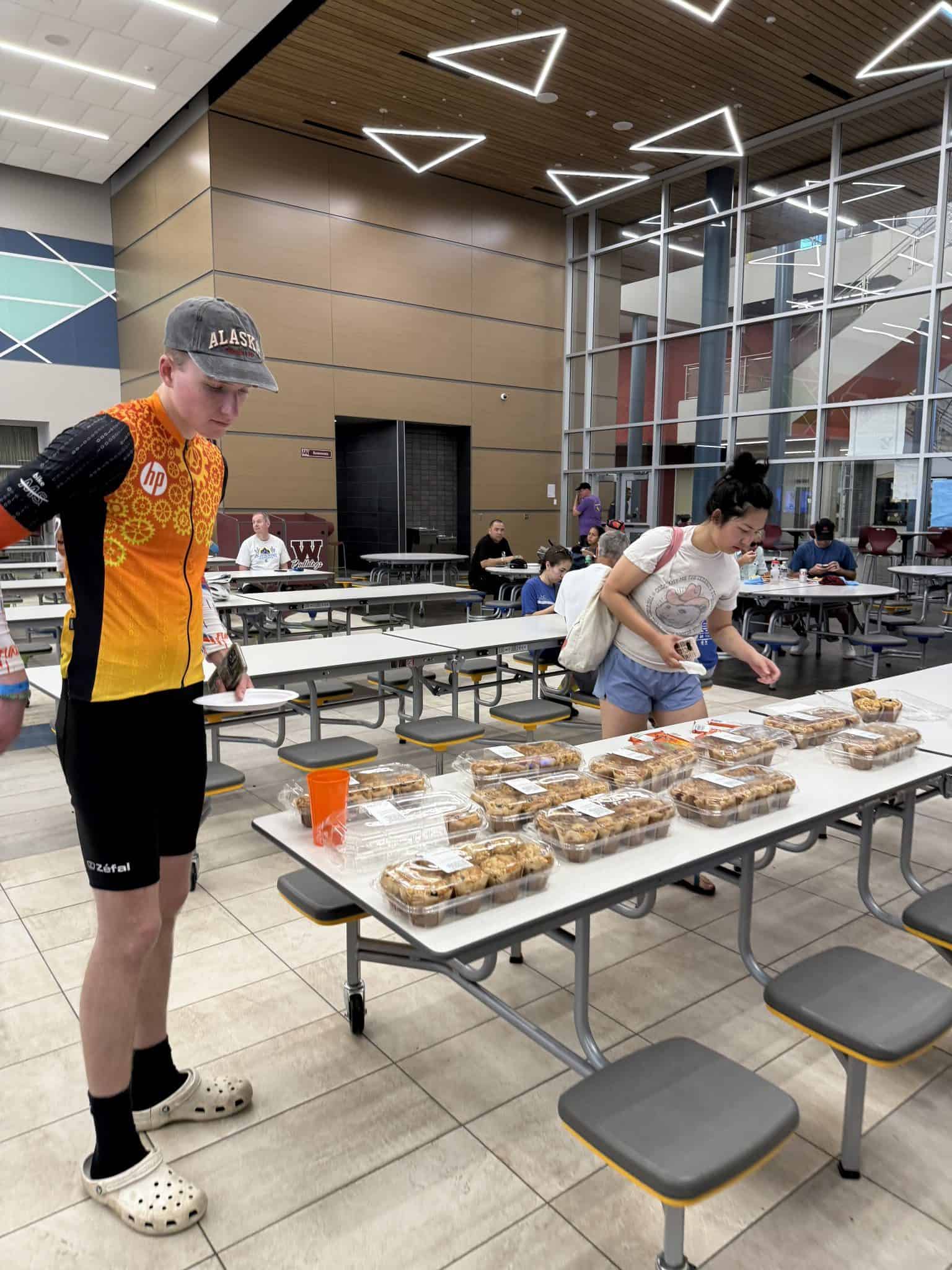 Bright indoor dining hall with young people browsing pre-packaged food trays. Modern architecture, high ceilings, geometric lighting.