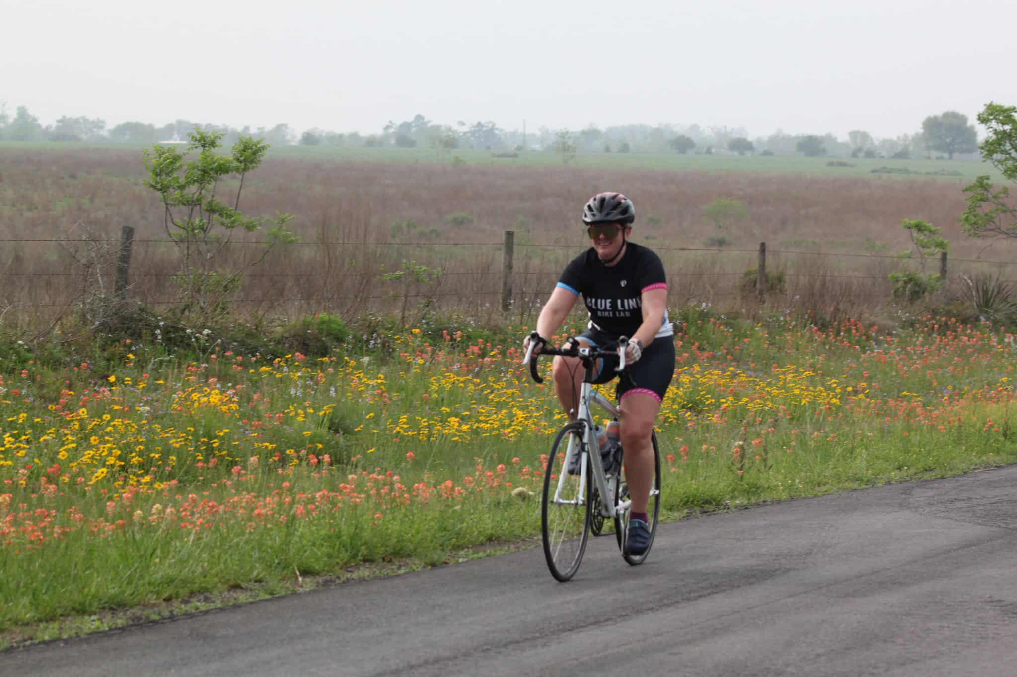 Cyclist riding on scenic country road with wildflowers and open fields, promoting Bluebonnet Express bike tours.