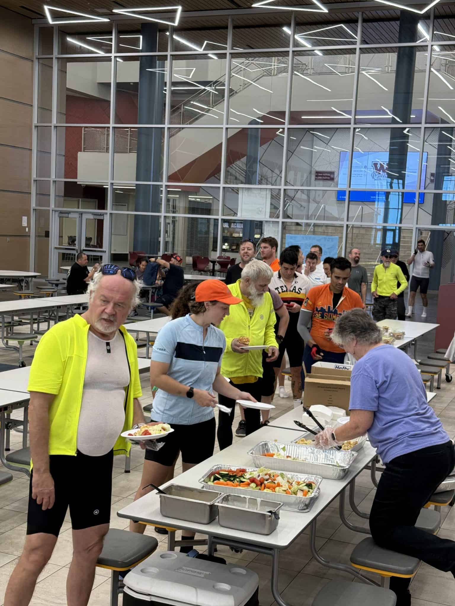 - Modern indoor space with large glass facade and people in cycling apparel at a food station.