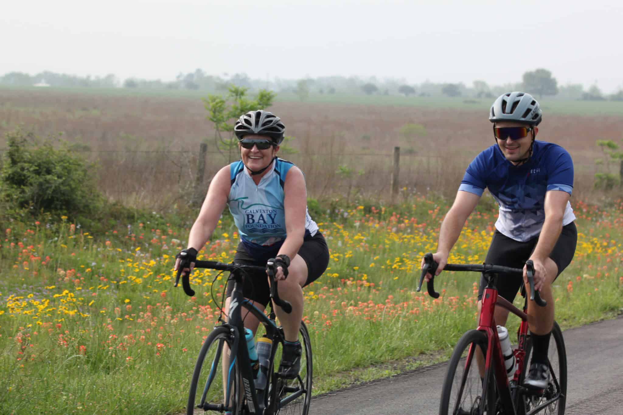Bicyclists riding on scenic countryside trail during daytime with wildflowers and open fields.