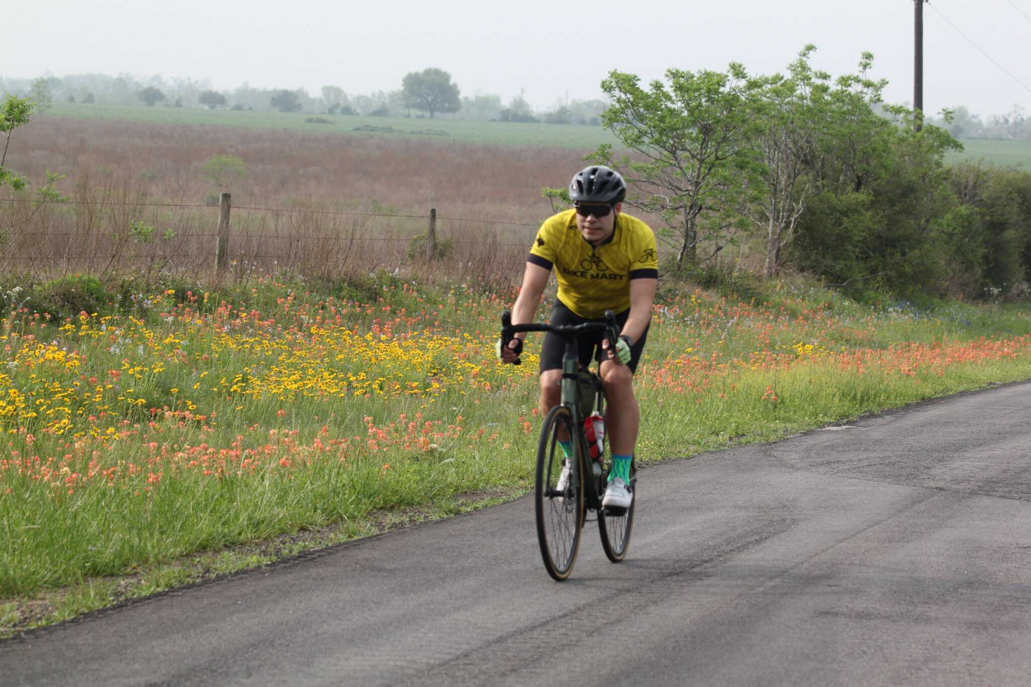 Bright yellow cyclist jersey on a road bike in scenic countryside with wildflowers and rolling hills.
