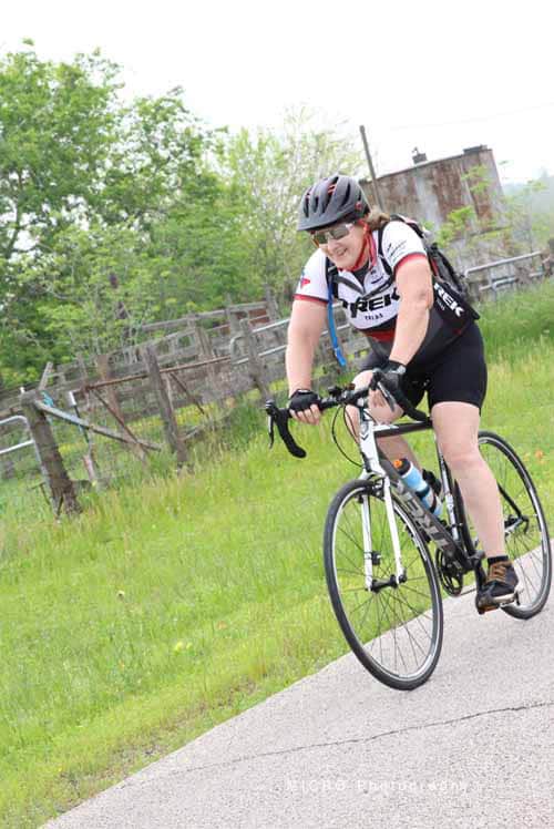 Vibrant cyclist riding on scenic trail with lush greenery and rustic fence, promoting Bluebonnet Express bike tours.