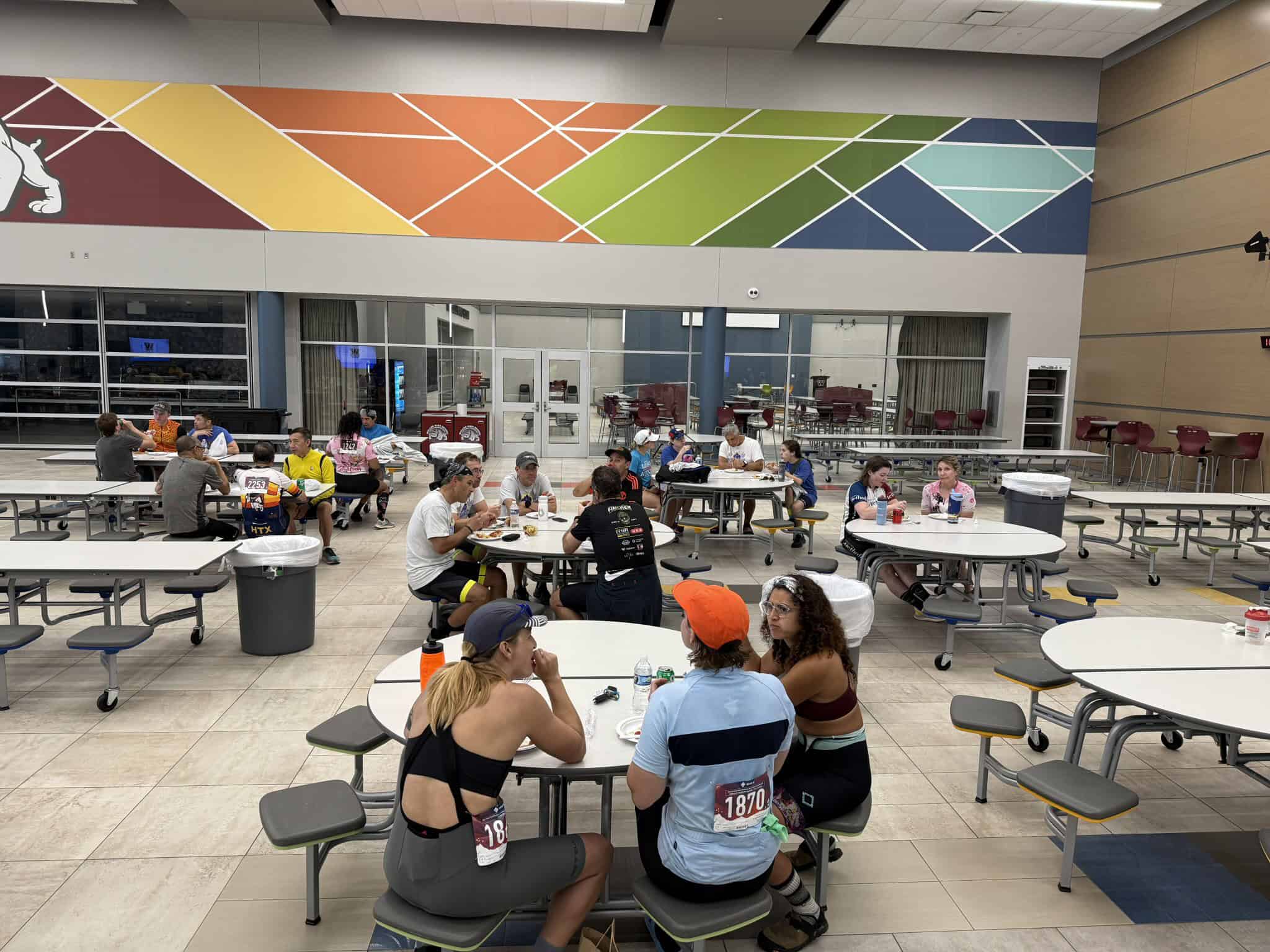 Bright indoor cafe at a cycling event featuring people eating and socializing, vibrant mural decor on the wall.