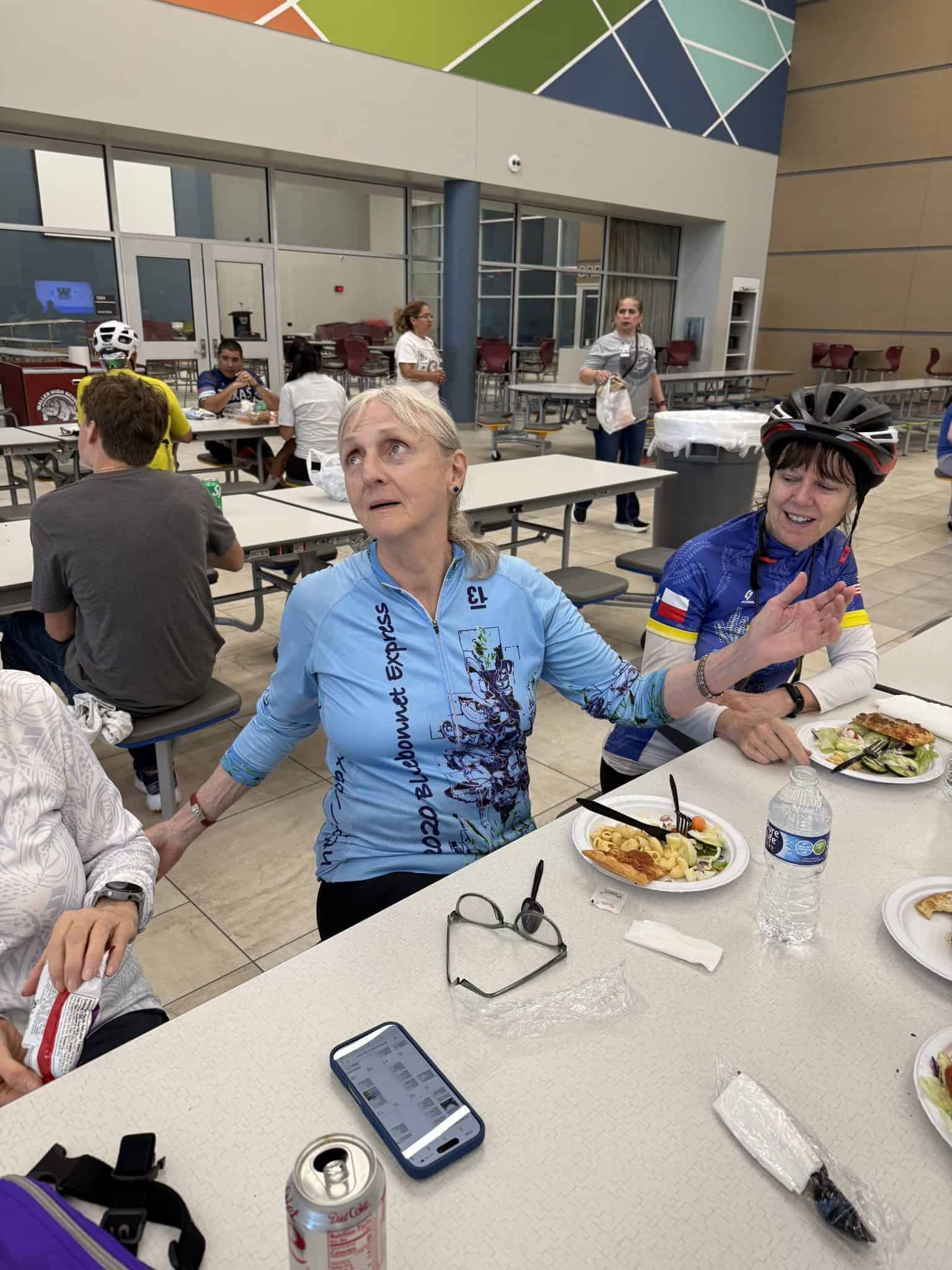 Older woman in blue cycling jersey at Bluebonnet Express bike event, indoor dining, engaging with friends.