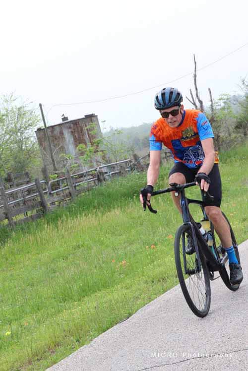 Bright cyclist riding on scenic trail with lush greenery and rustic fence in the background.