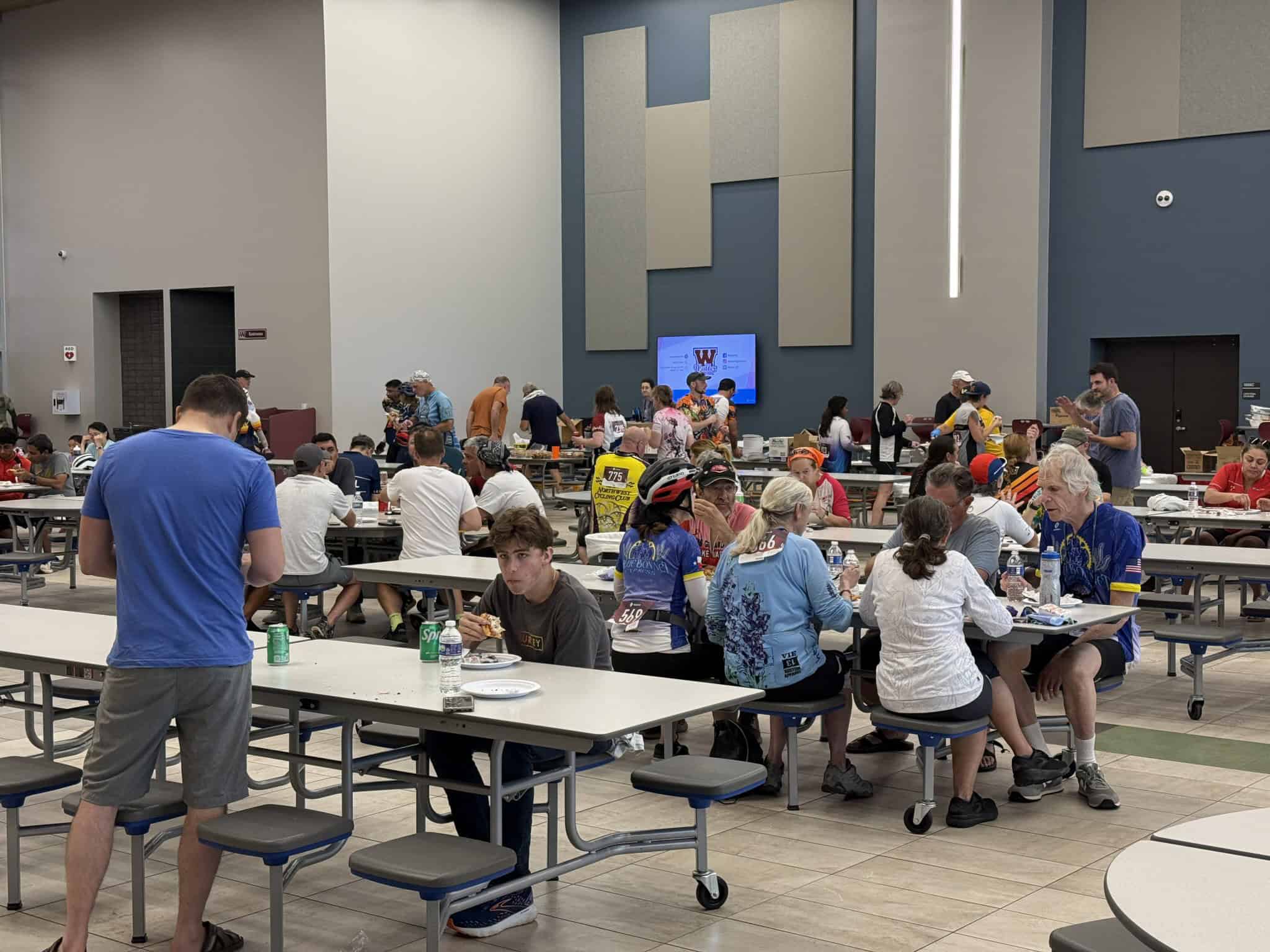 Bright indoor event space filled with cyclists and attendees enjoying meals at long tables during the Bluebonnet Express cycling event.