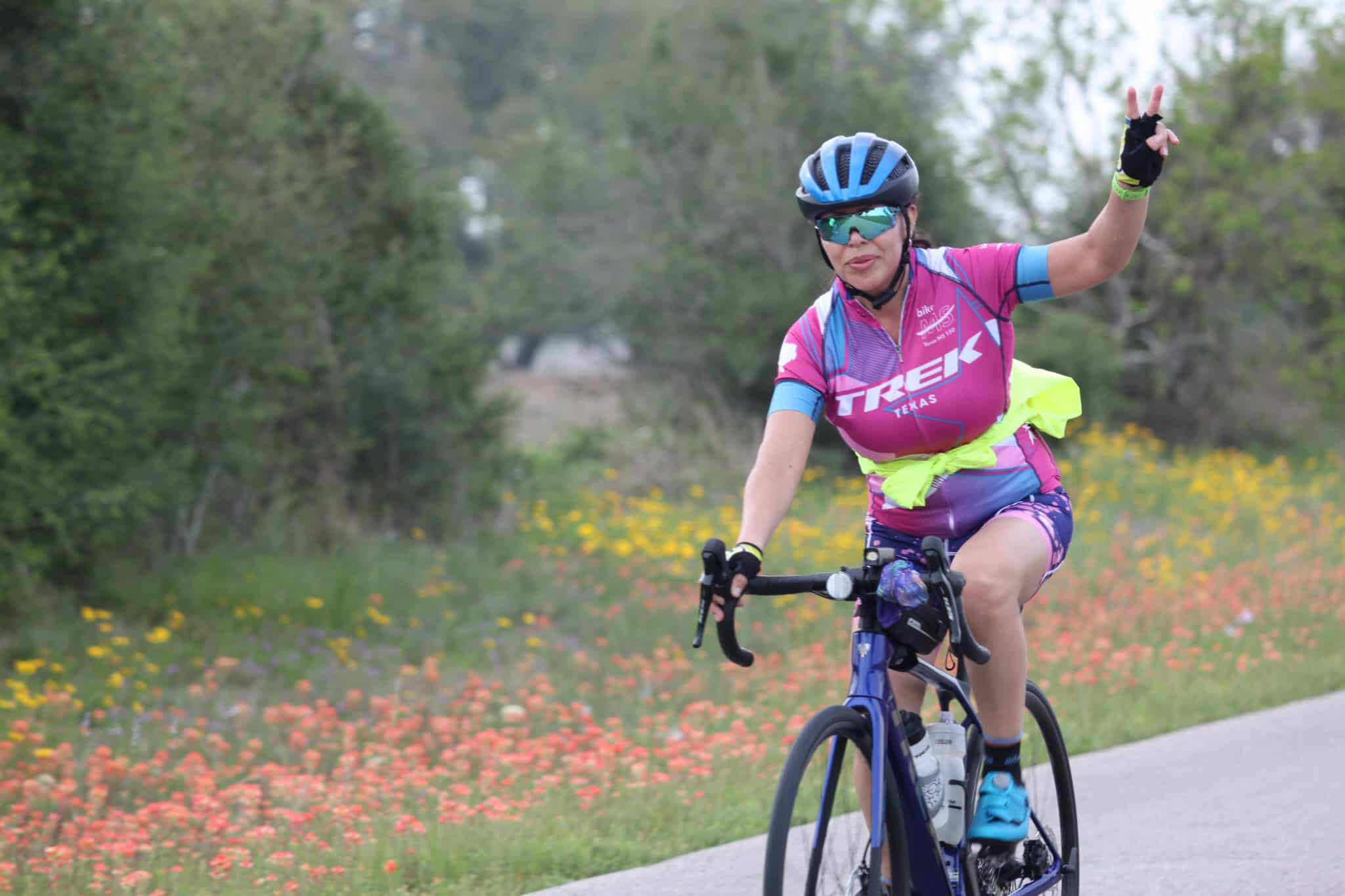 Bright female cyclist in Trek Texas gear making peace sign on scenic bike trail.