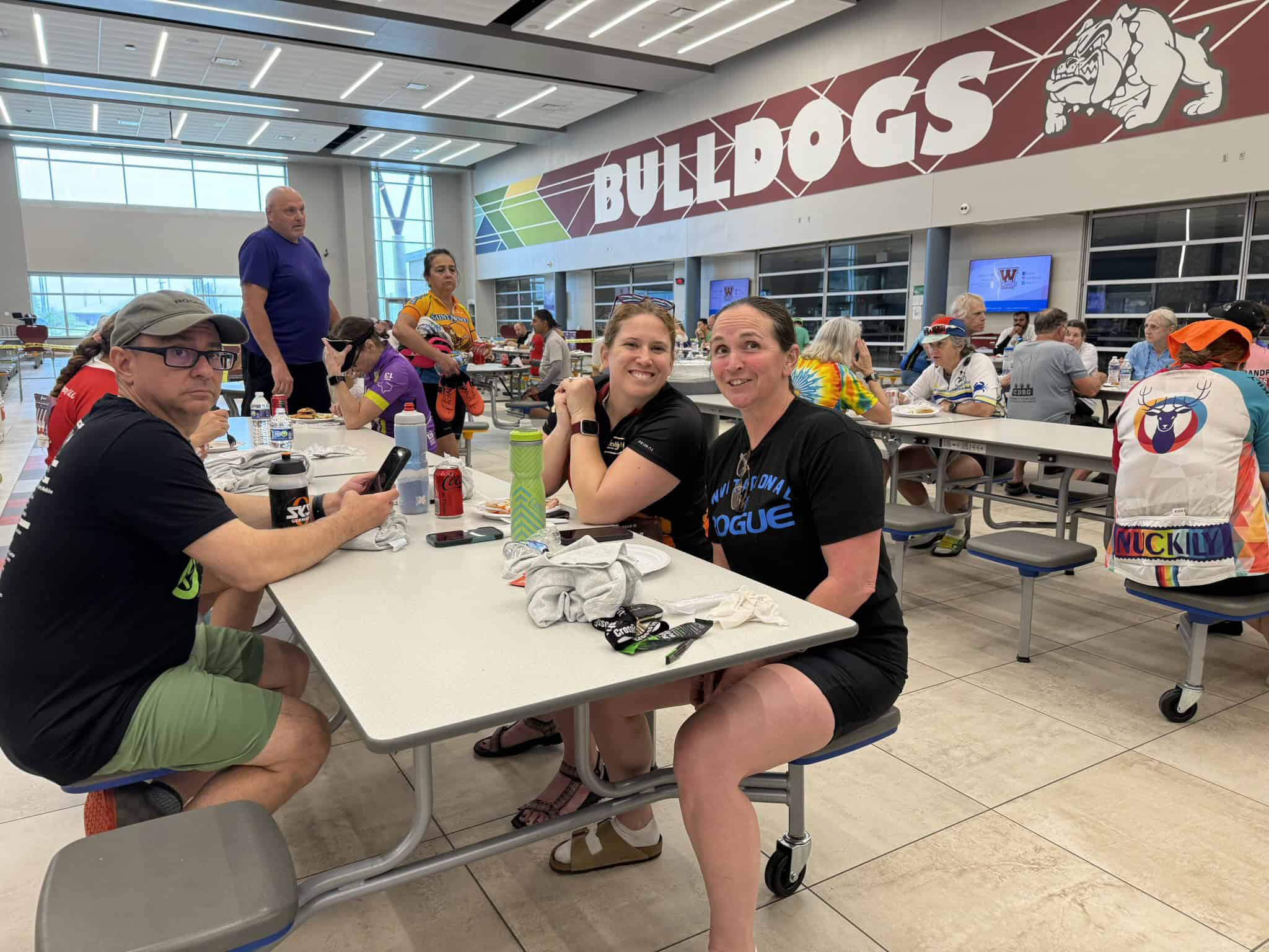 Bright indoor cafe at a sports facility with cyclists and supporters, featuring "Bulldogs" branding and a lively atmosphere.