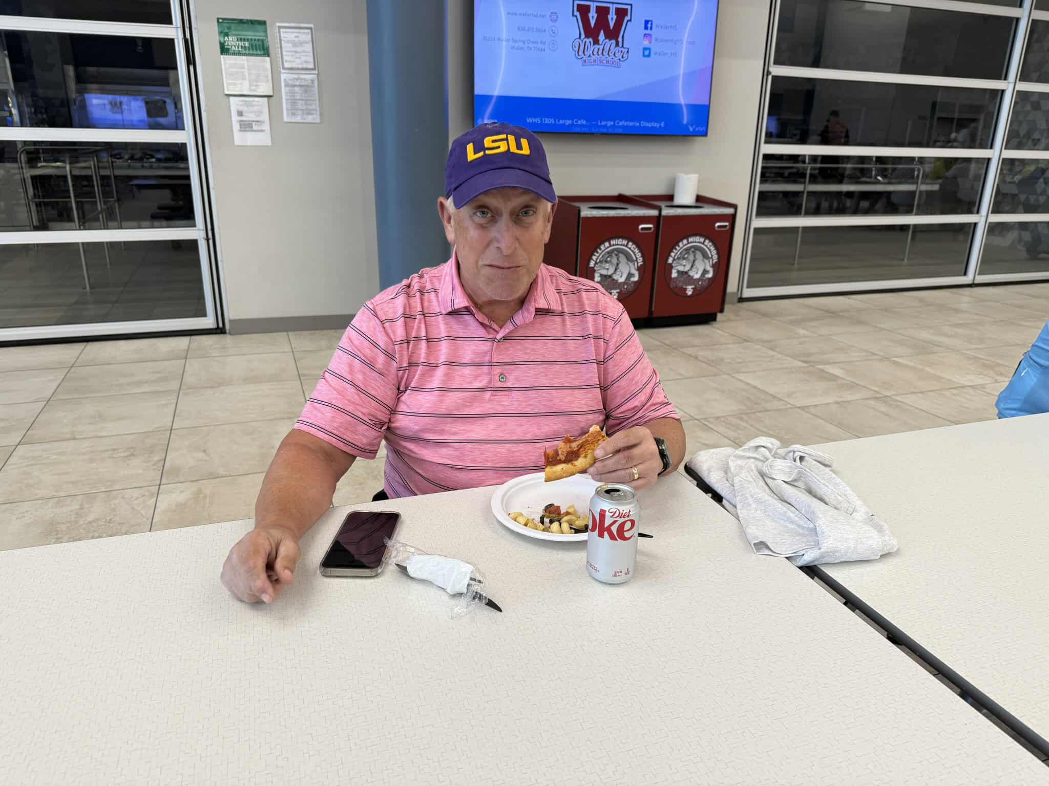 Lawn care worker resting at school cafeteria, eating pizza, with Diet Coke and phone on table.