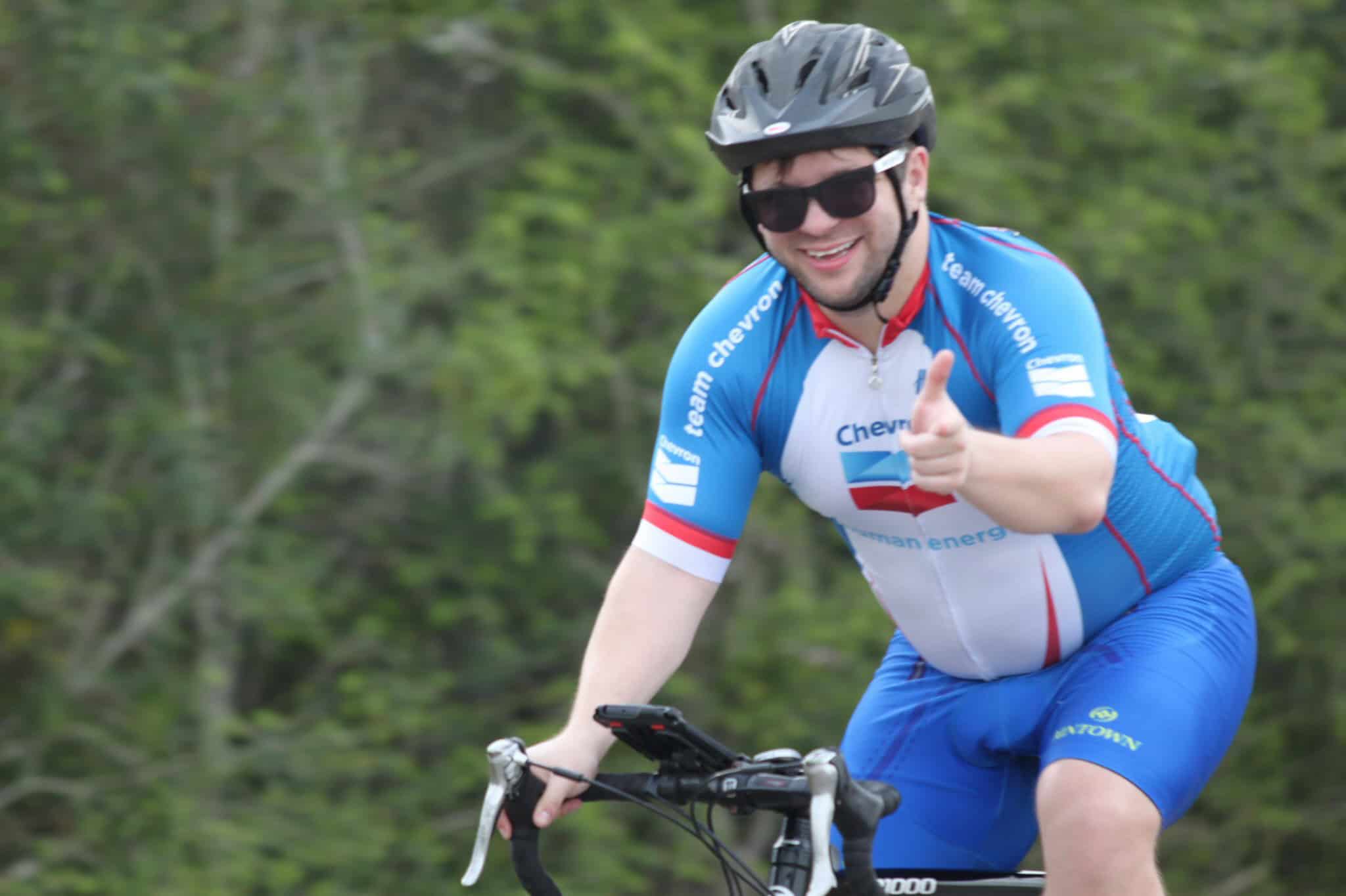 Bright male cyclist in blue Chevrón team jersey smiling, pointing, and wearing a helmet during outdoor ride.
