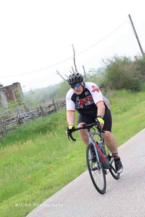 Helmeted cyclist riding on a rural road with green fields and a rustic barn in the background.