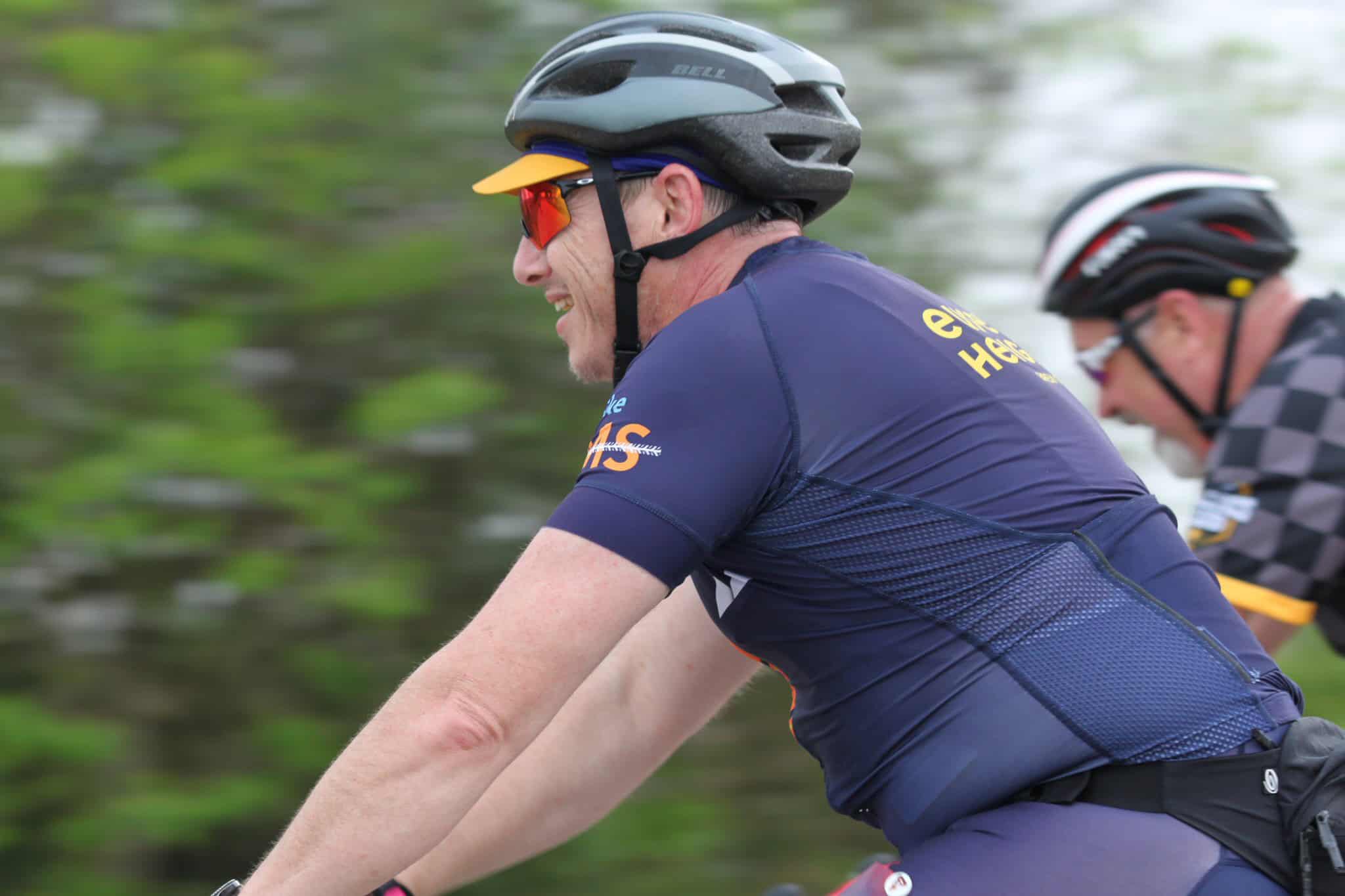 Bright-eyed cyclist wearing a blue jersey, helmet, and sunglasses riding outdoors, promoting Bluebonnet Express bike tour.