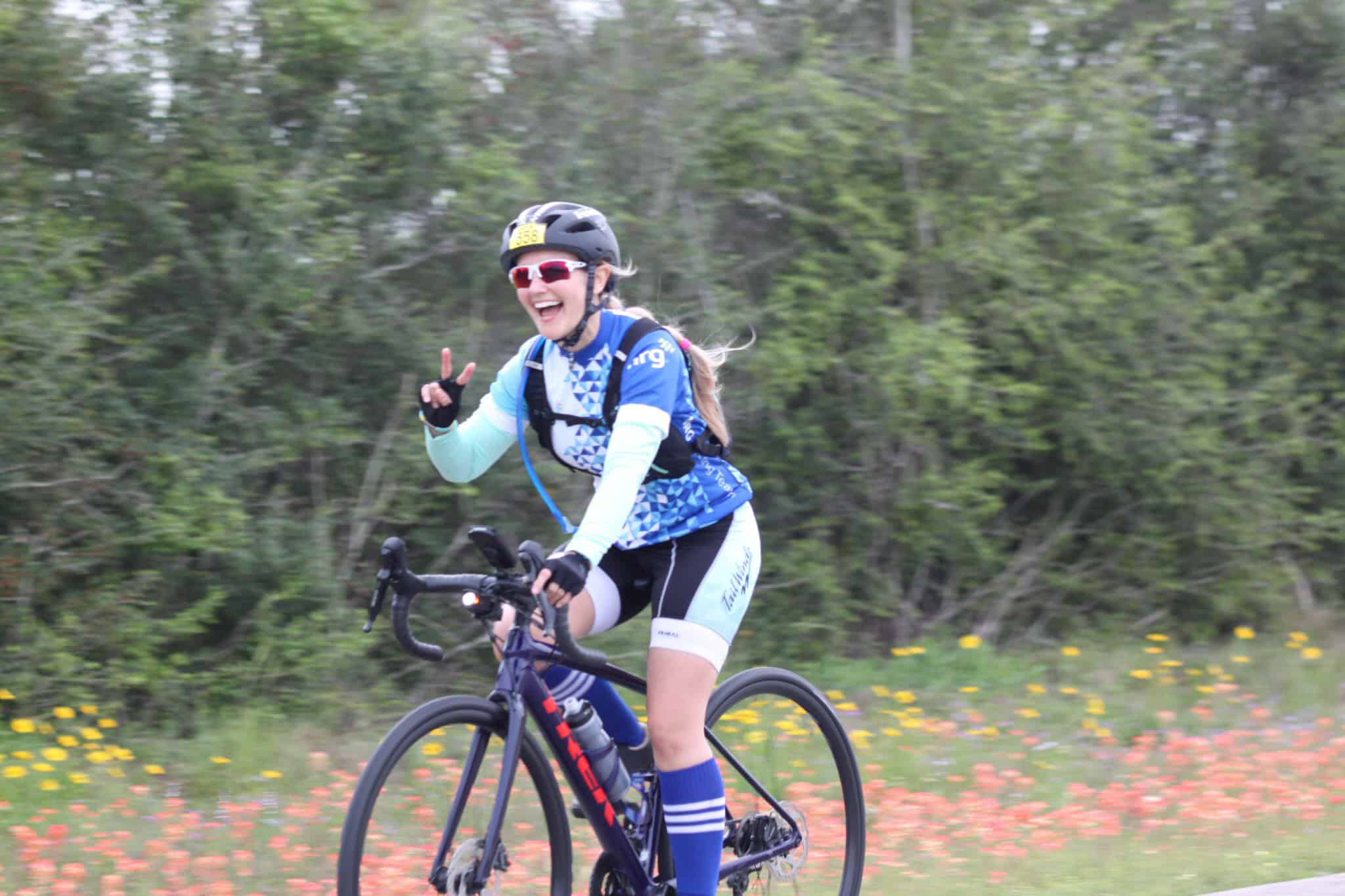 Vibrant woman cycling on scenic trail, smiling and making peace sign, outdoor adventure with Bluebonnet Express.