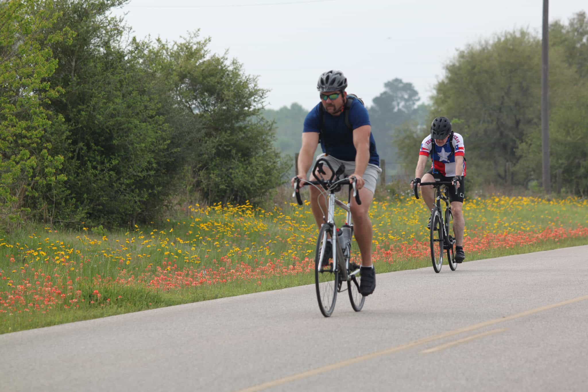 Riders cycling on scenic country road with wildflowers, promoting Bluebonnet Express bike tours.