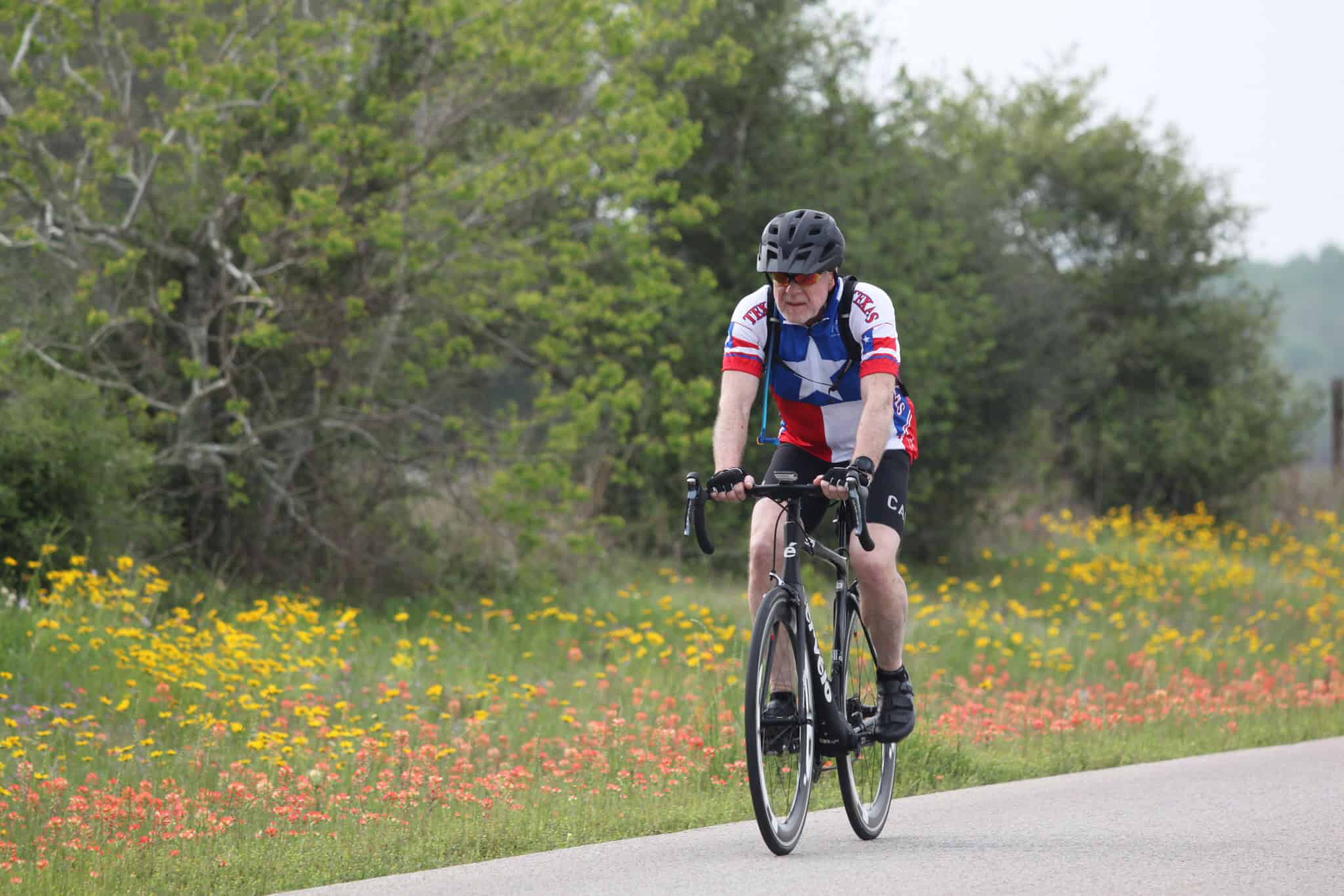 Colorful cyclist riding on scenic countryside road with wildflowers and green trees, promoting Bluebonnet Express bike tours.