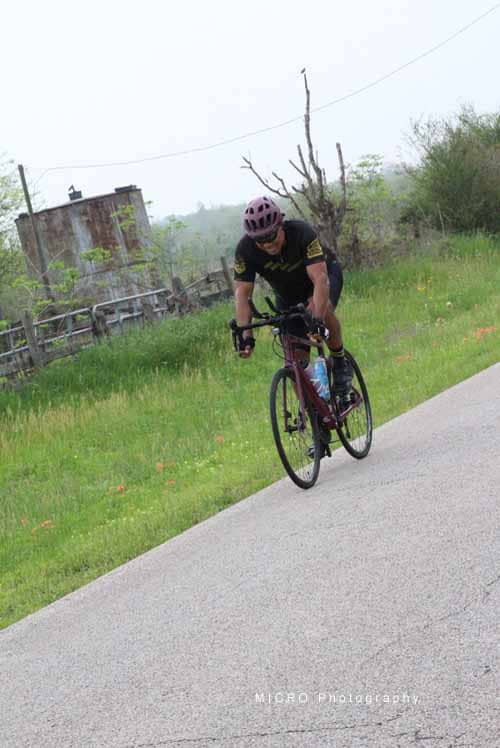 Bicycle rider on rural road with green landscape and rustic building in background.