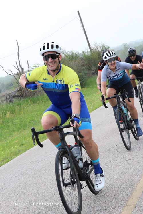 Smiling cyclist wearing blue and yellow jersey riding along rural road during Bluebonnet Express event.