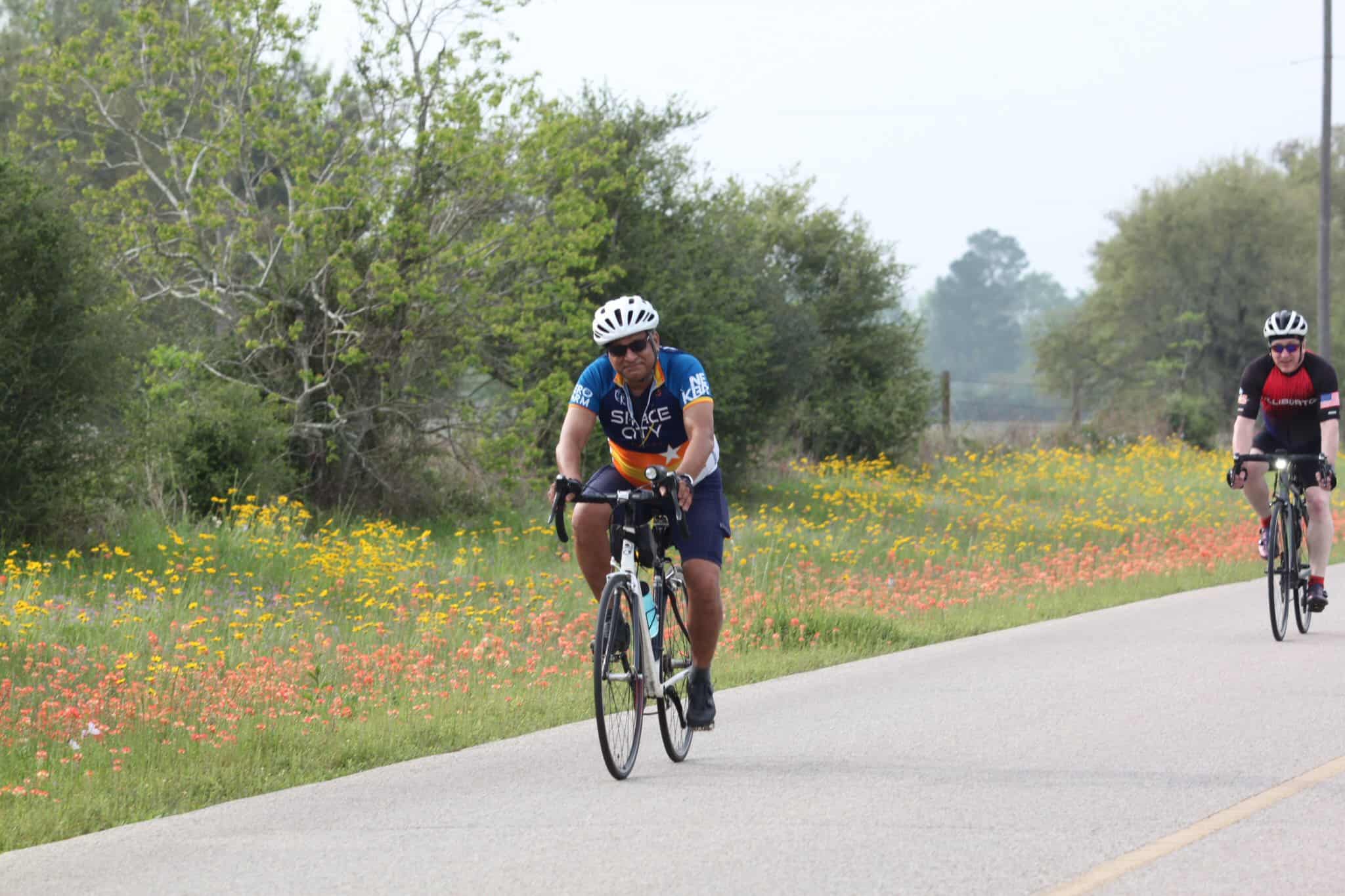 Bright blue and black cycling jerseys on two cyclists riding through a scenic countryside route with colorful wildflowers.