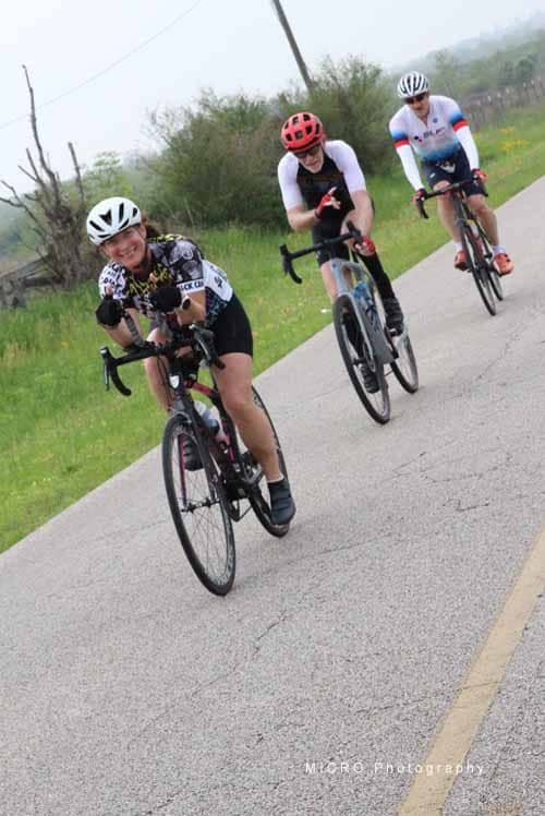 Smiling cyclists riding on a rural road during daytime, promoting Bluebonnet Express cycling event.