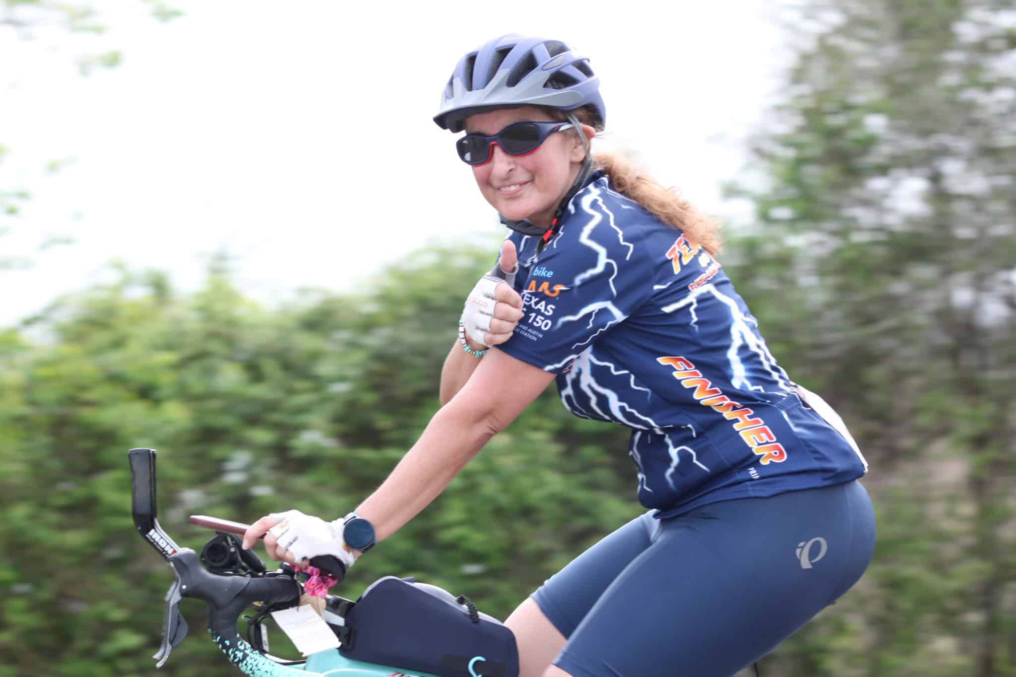 Active woman cyclist wearing Bluebonnet Express jersey riding bike outdoors, smiling with thumbs up.