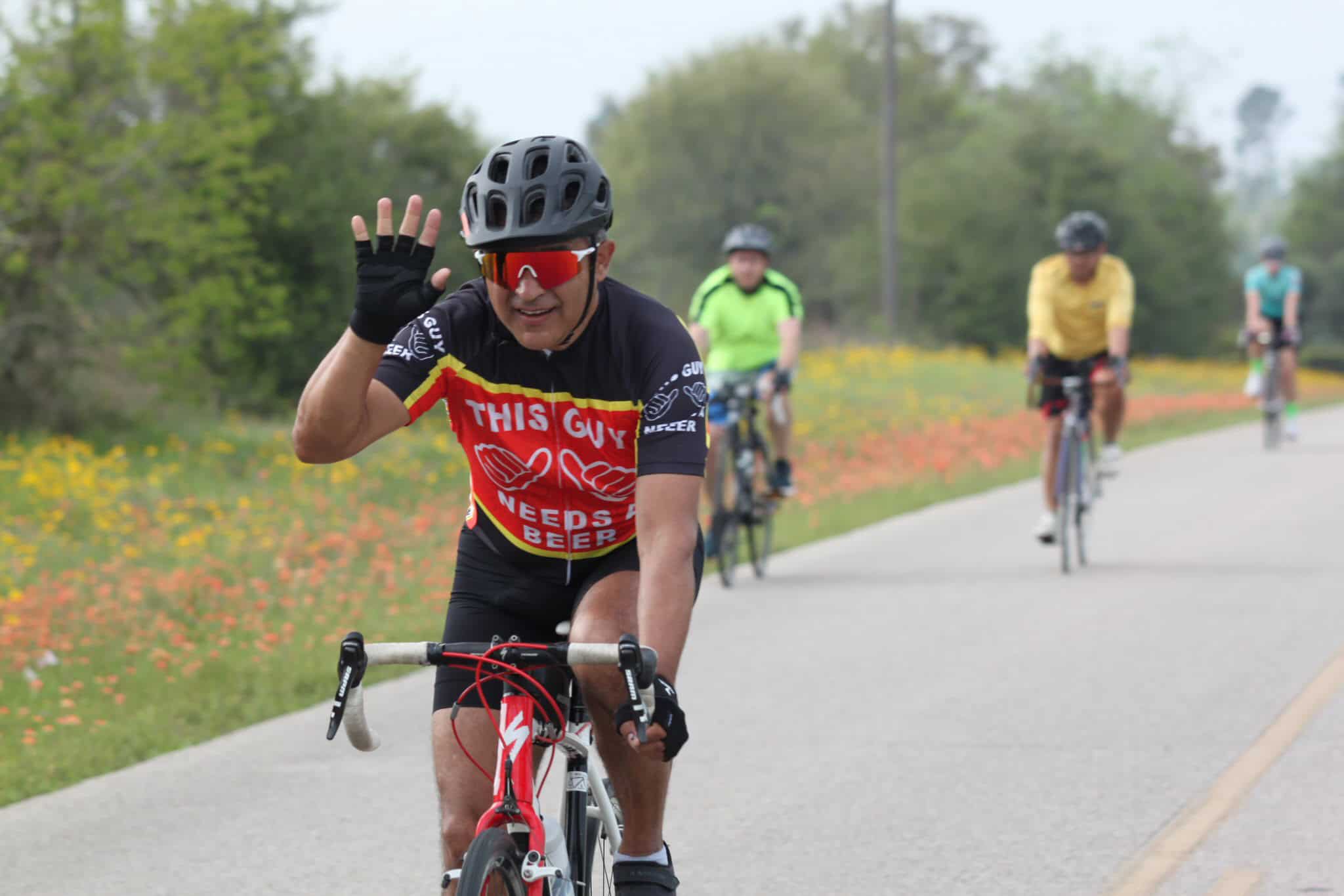 Bicycle rider waving during a scenic outdoor ride with fellow cyclists on a sunny day.