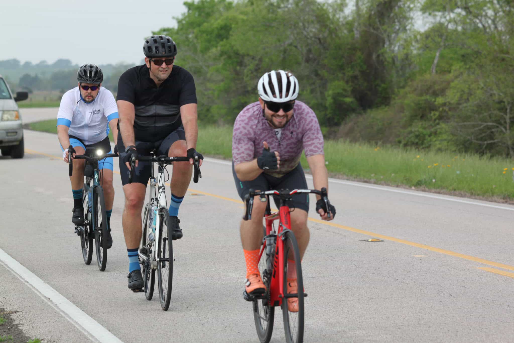 Vibrant group cycling on scenic road with lush green trees, showcasing Bluebonnet Express bicycle adventure.