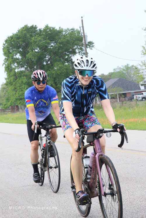 Bright female cyclist wearing a blue jersey and colorful shorts riding a pink bike on a suburban street.