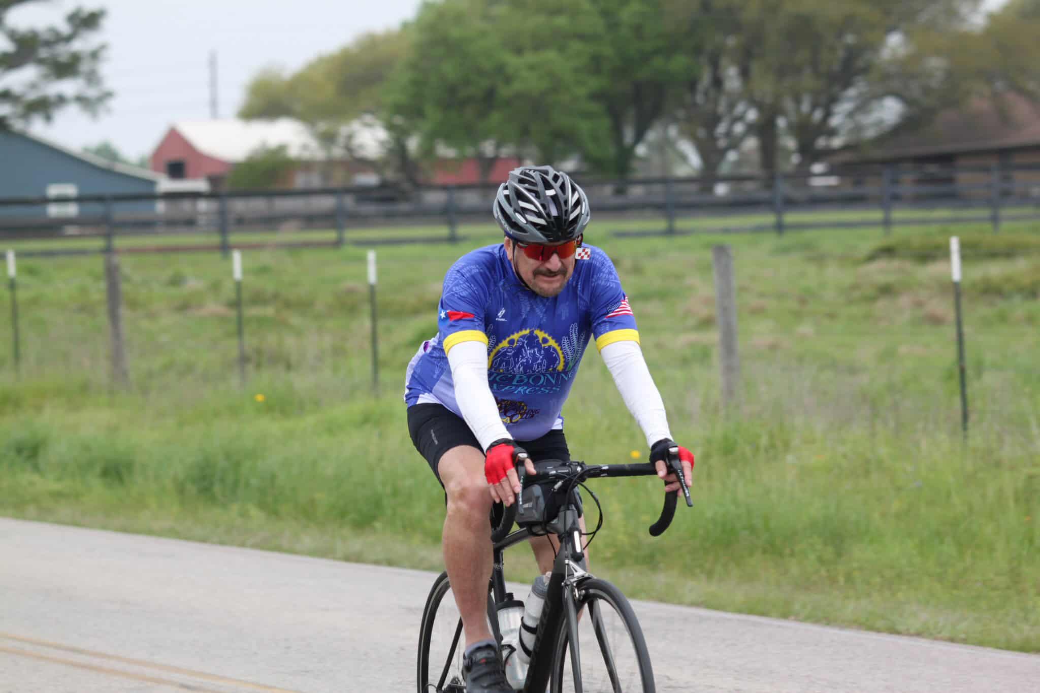 Bright blue cyclist jersey with "Bluebonnet Express" logo on a road bike, riding through a scenic rural area.