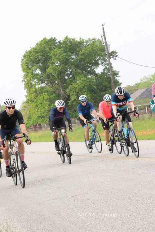 Bright group of cyclists riding on a rural road during daytime, promoting Bluebonnet Express bike event.