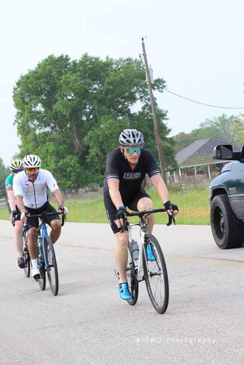 Bike riders in Bluebonnet Express cycling event on rural road with trees and houses in background.
