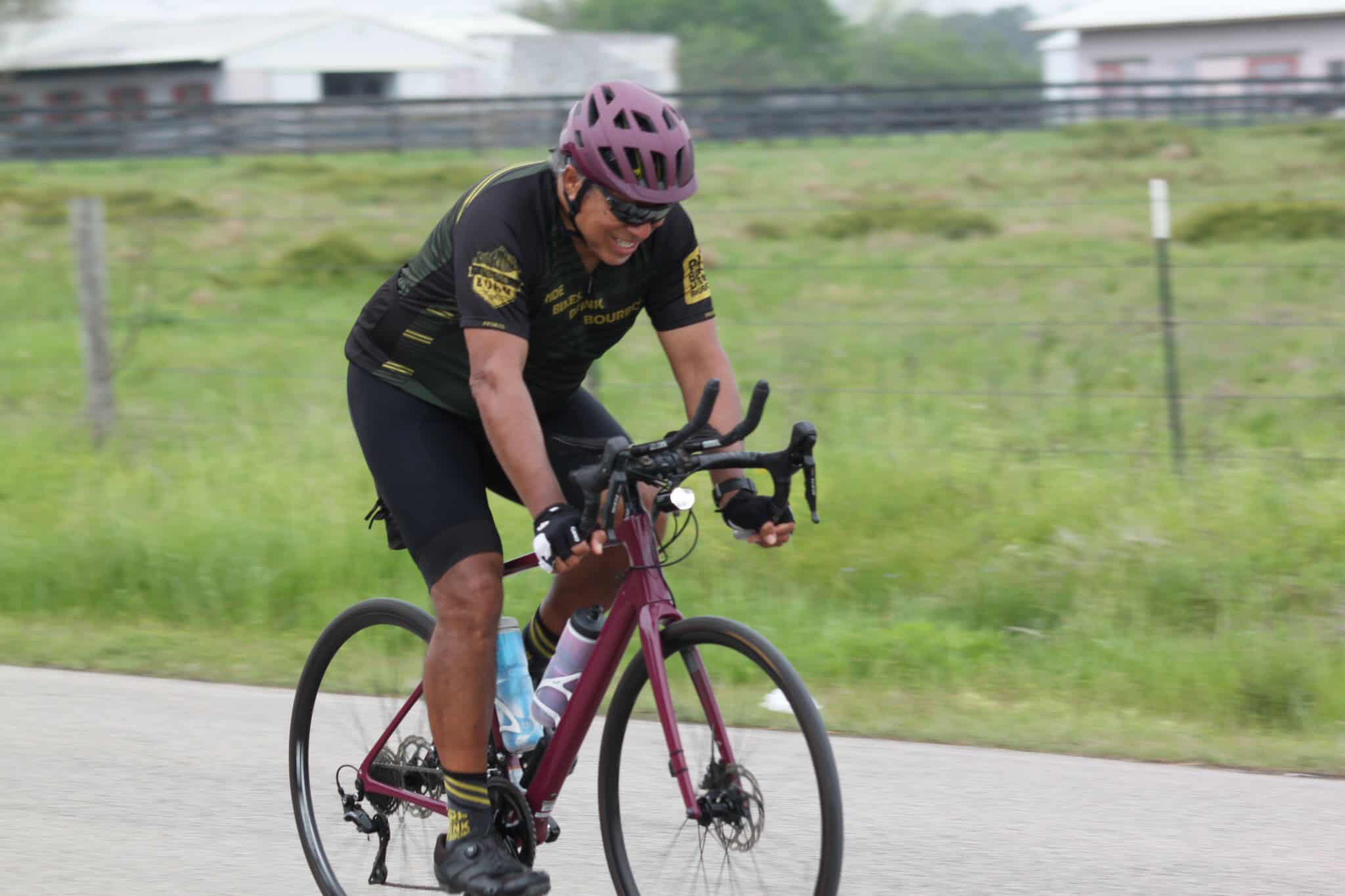 Vibrant cyclist riding a bike on a scenic trail with green fields and a purple helmet.