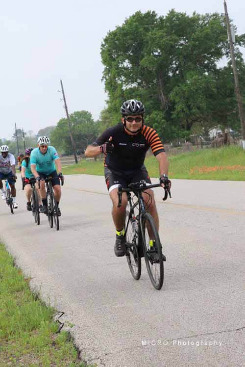 Bright cyclist wearing black and orange jersey leading a group along a rural road during Bluebonnet Express event.