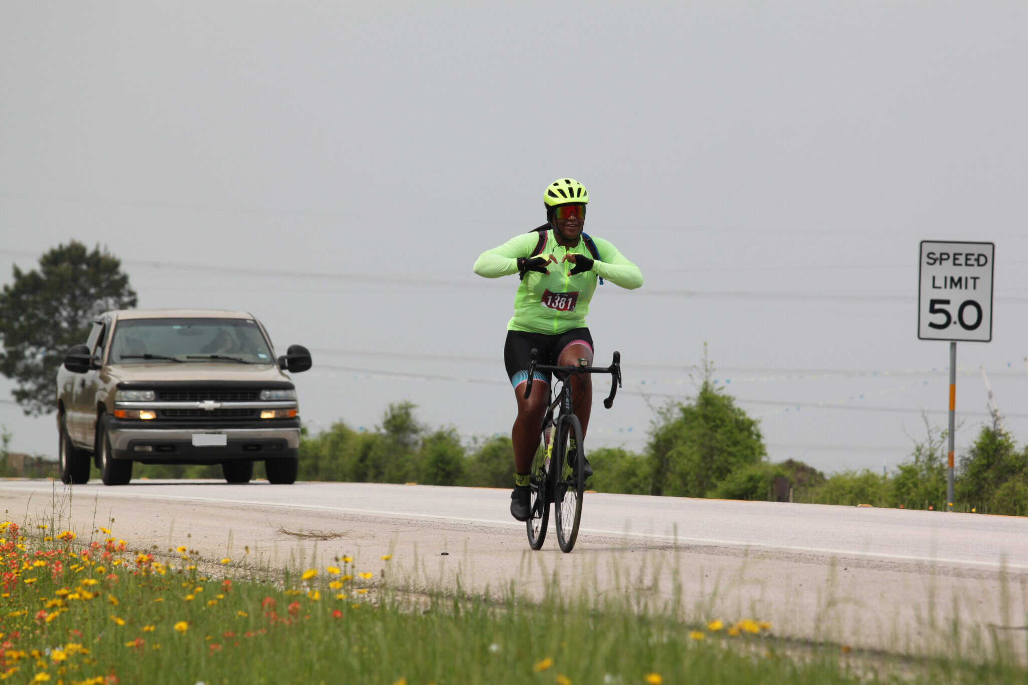 Cyclist riding on open road during race, wearing bright neon gear, with vehicle and speed limit sign background.